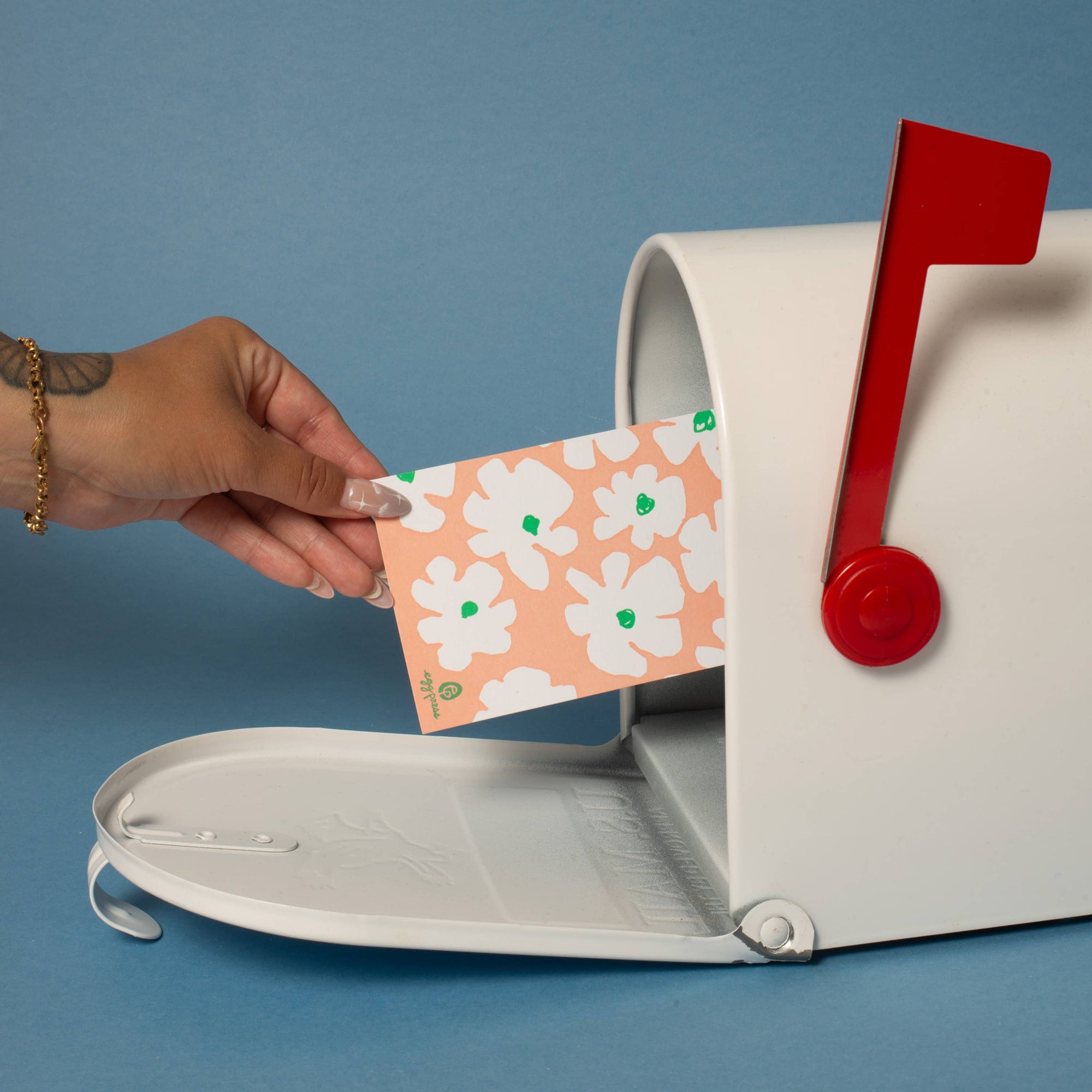 Hand placing a card with floral design into a mailbox against a blue background