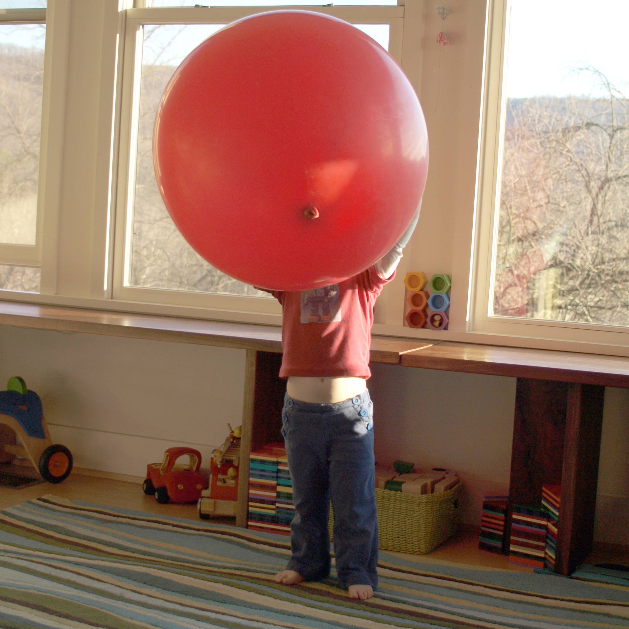 A child holding a large red balloon in front of a window, with toys on the floor.