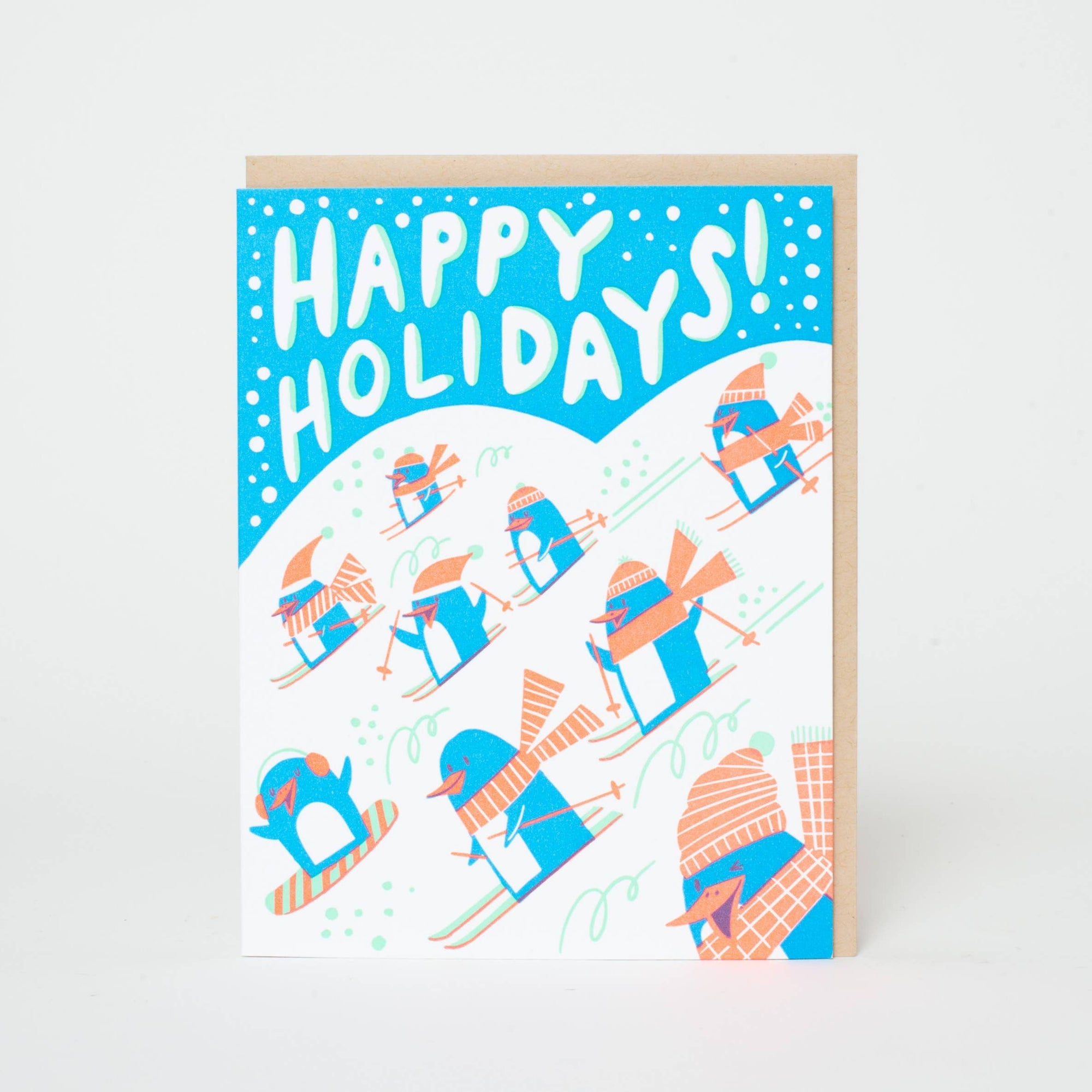 A greeting card with a hand-letterpress printed 'Happy Holidays!' message, featuring cartoon penguins in a wintry landscape, accompanied by a kraft envelope.