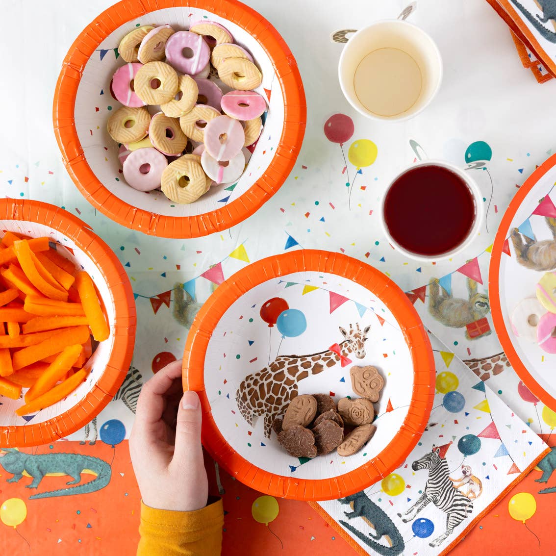 Children's party table with paper plates, snacks, and drinks on a colorful tablecloth.