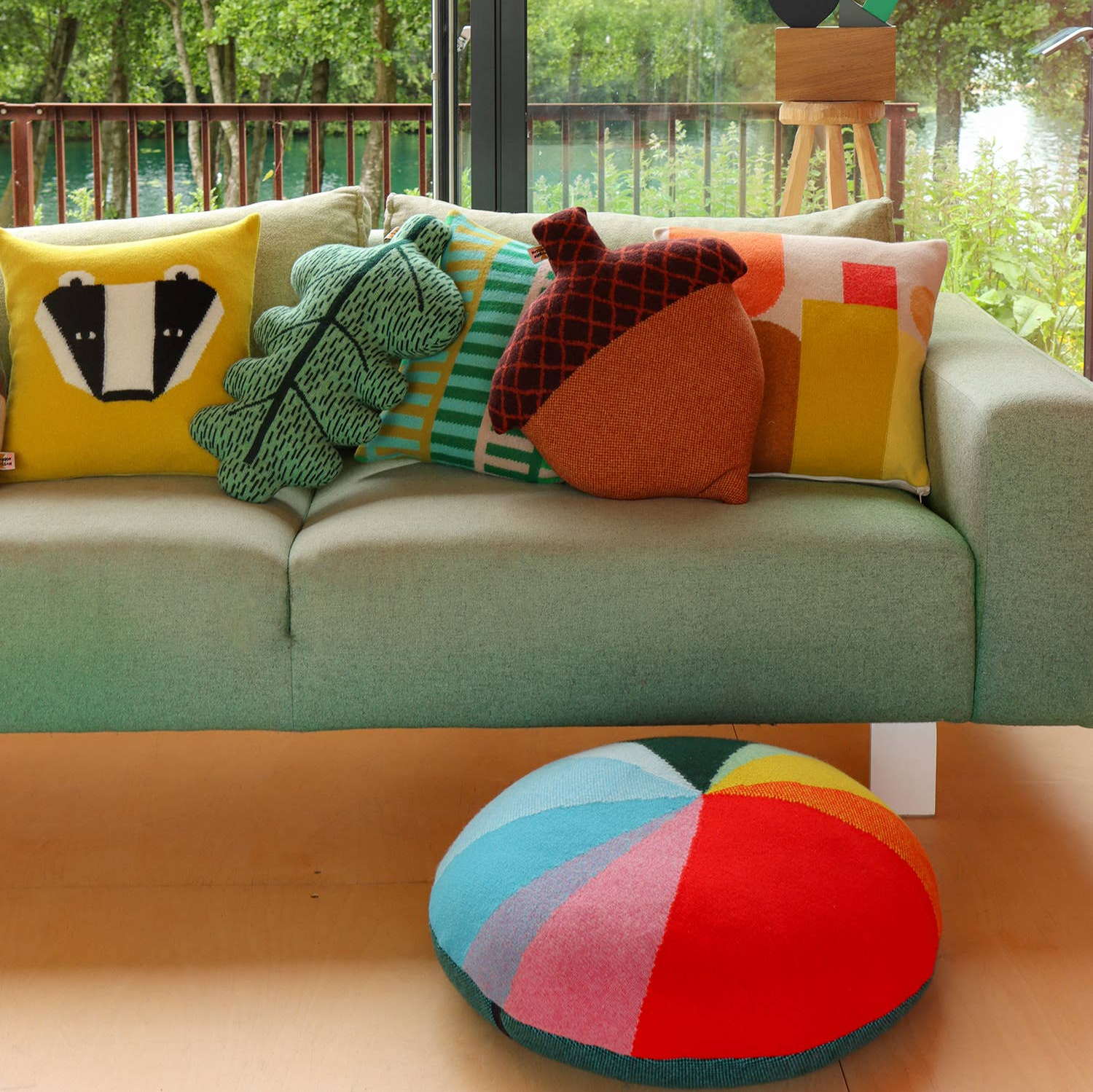 A colorful knitted floor cushion with geometric patterns placed on a sofa alongside other cushions.