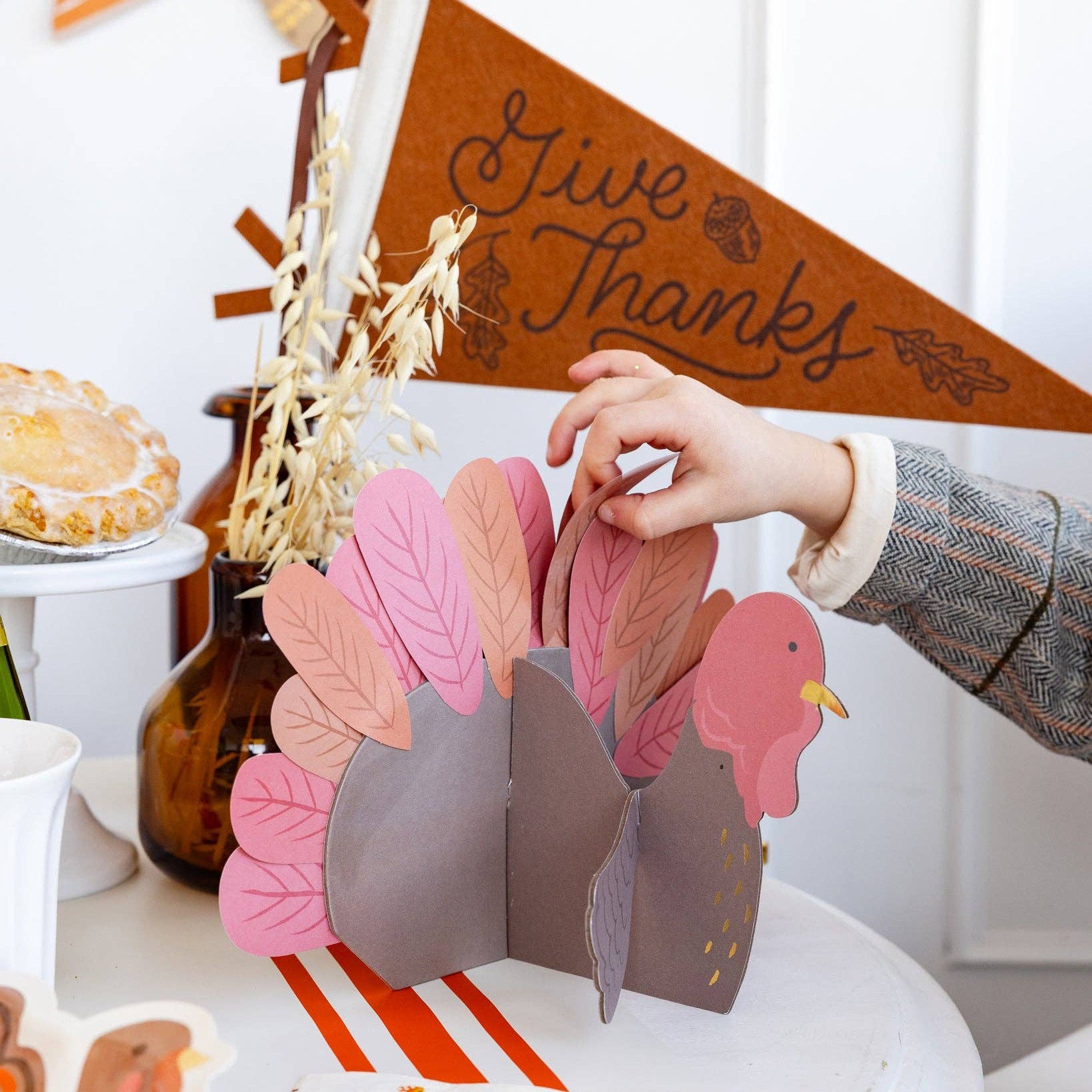 Children's hand interacting with a paper turkey craft on a table with a 'Give Thanks' banner in the background.