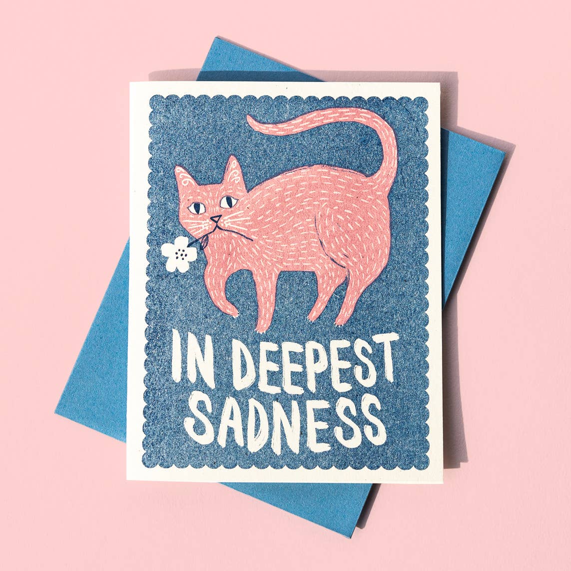 A greeting card with an illustration of a cat holding a flower, with the text 'IN DEEPEST SADNESS' on it. The card is placed against a pink background, and a blue envelope is partially visible to the side.