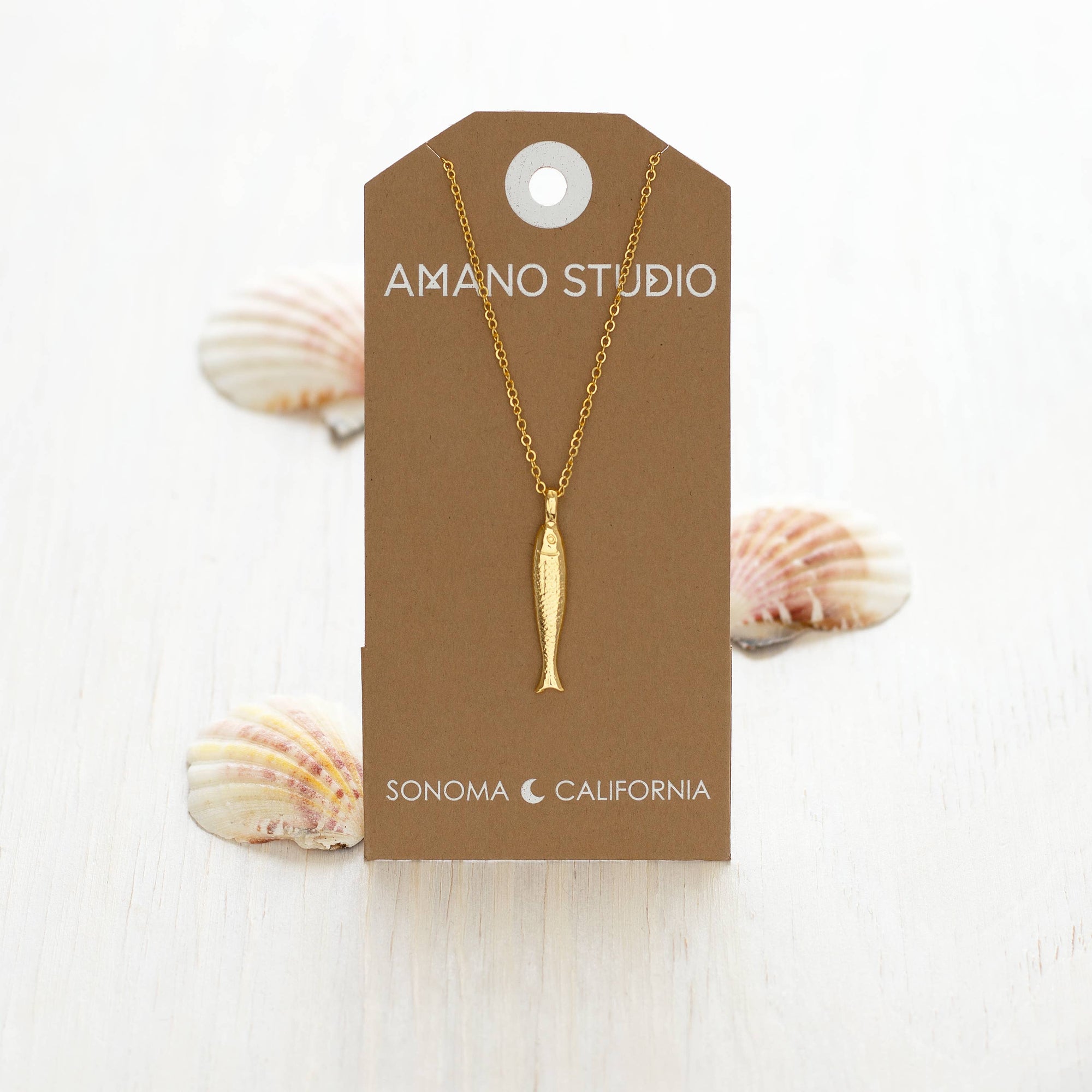Gold fish pendant necklace on a brown AMANO STUDIO card with seashells in the background.