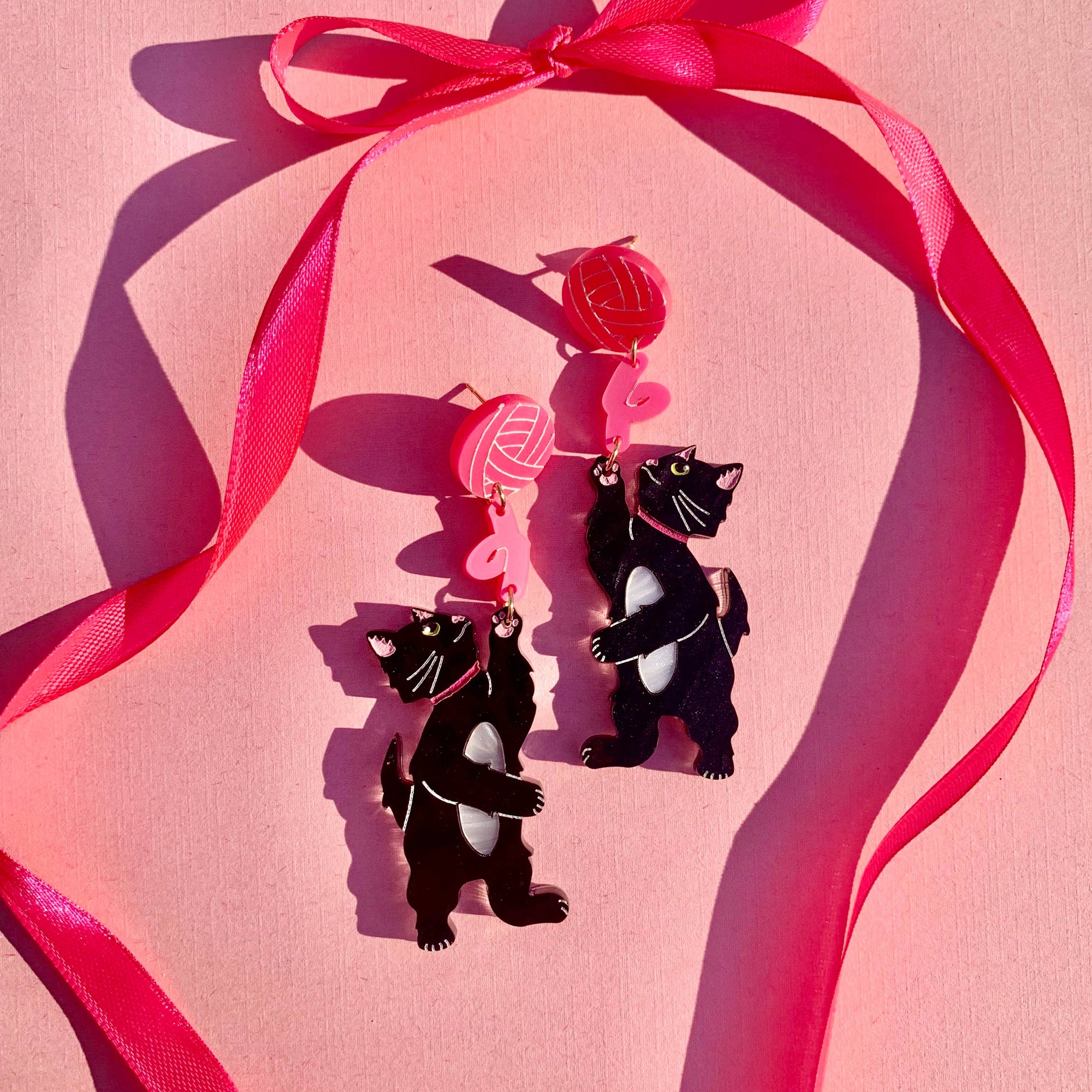 Black cat earrings with pink ribbons on a pink background