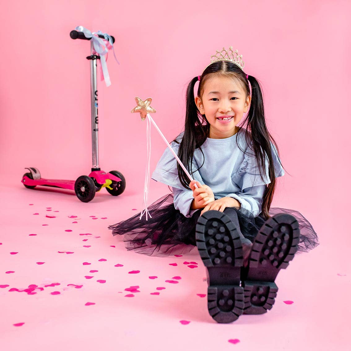 Child with a tiara and wand on a pink background with a scooter.