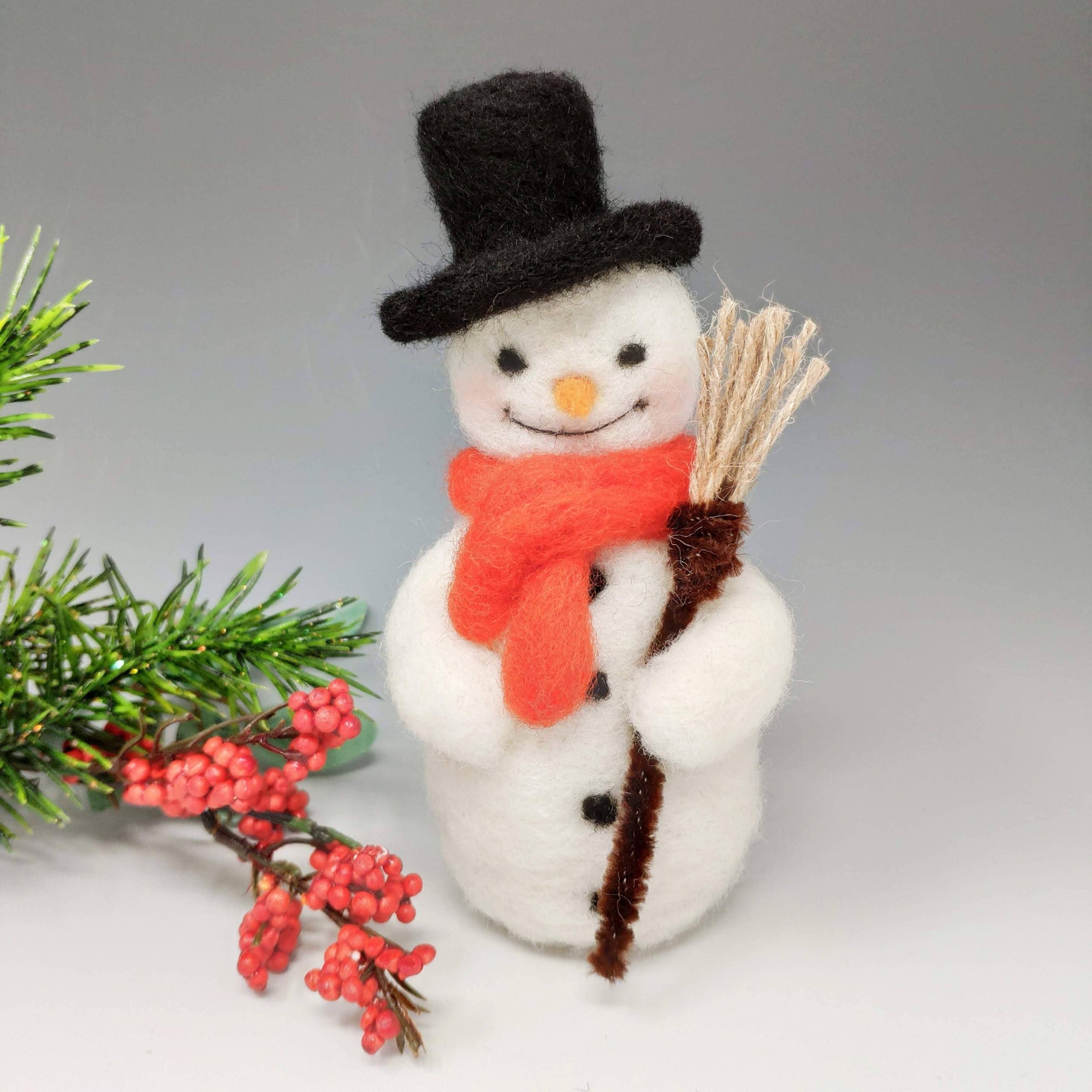 A needle felted snowman craft kit with a black hat, orange scarf, and holding a broom, displayed next to festive berries and greenery.