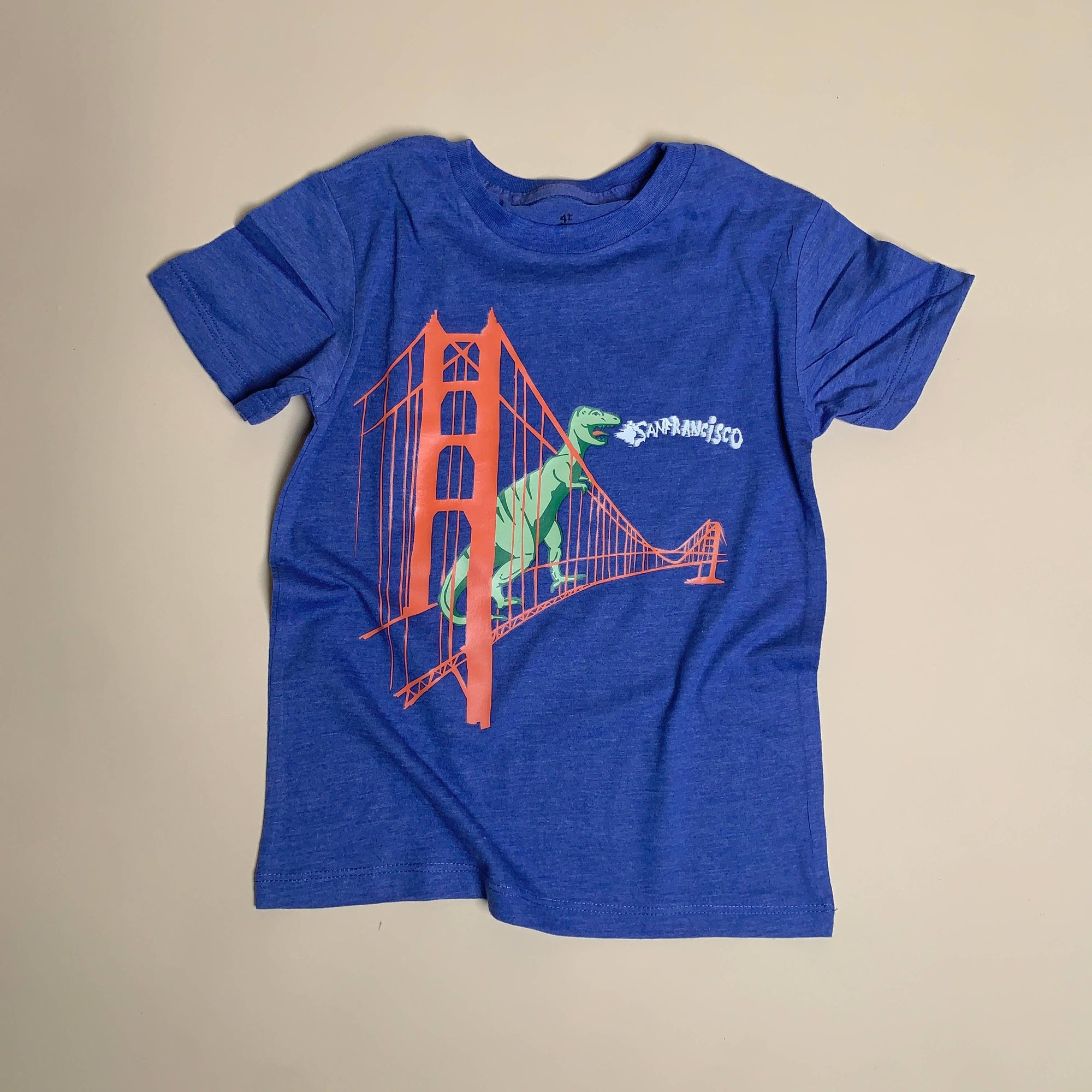 Blue t-shirt with a graphic of a dinosaur crossing the Golden Gate Bridge on a beige background