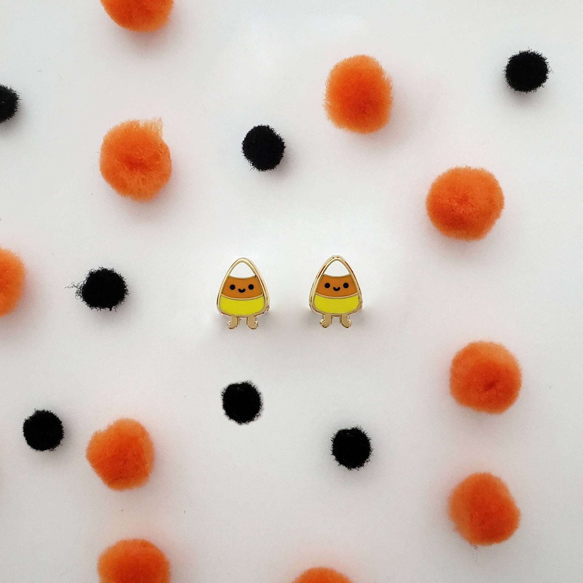 Pair of candy corn-shaped earrings on a white background with orange and black pom-poms.