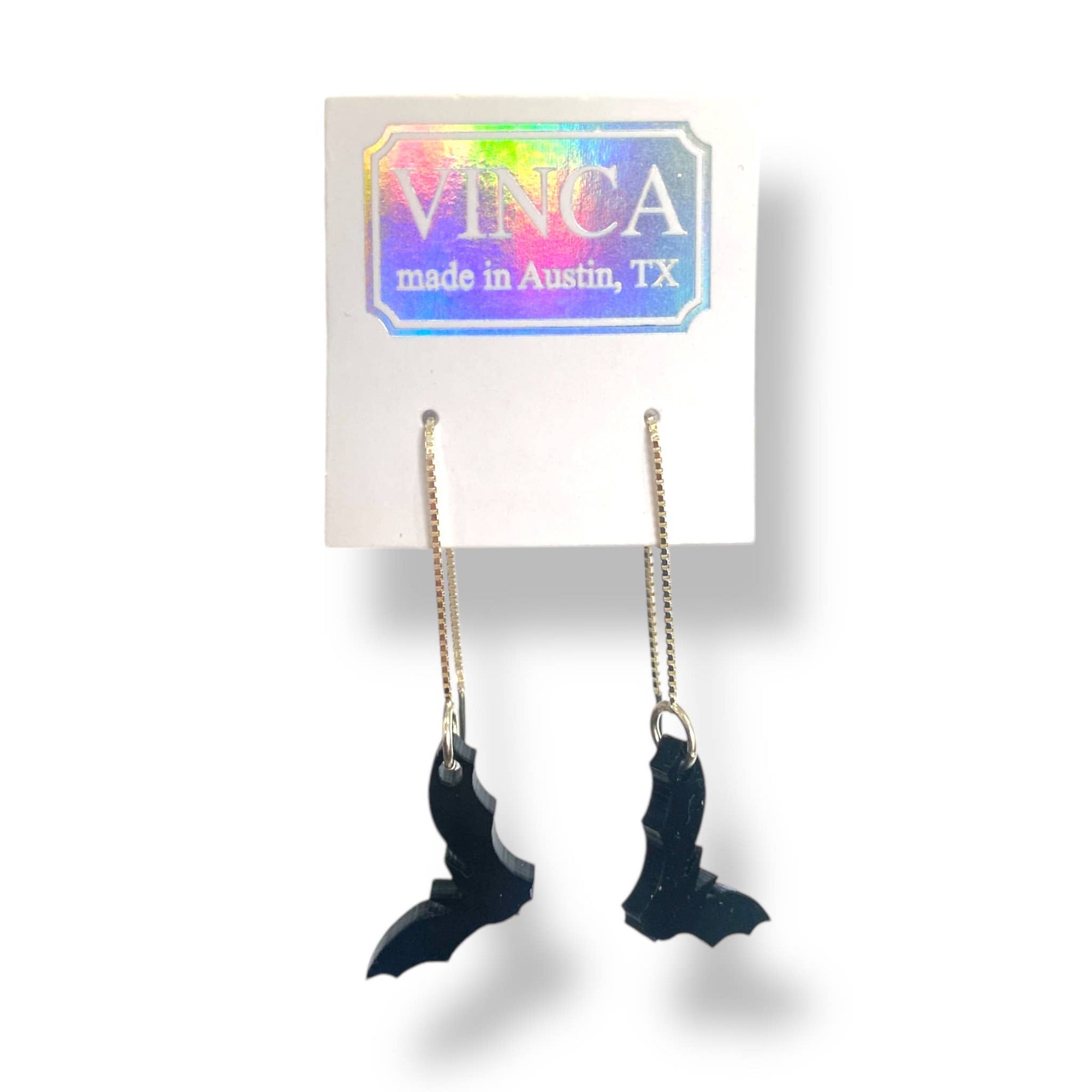 Black wing-shaped earrings on gold chains with a 'VINCA' branded card in the background.