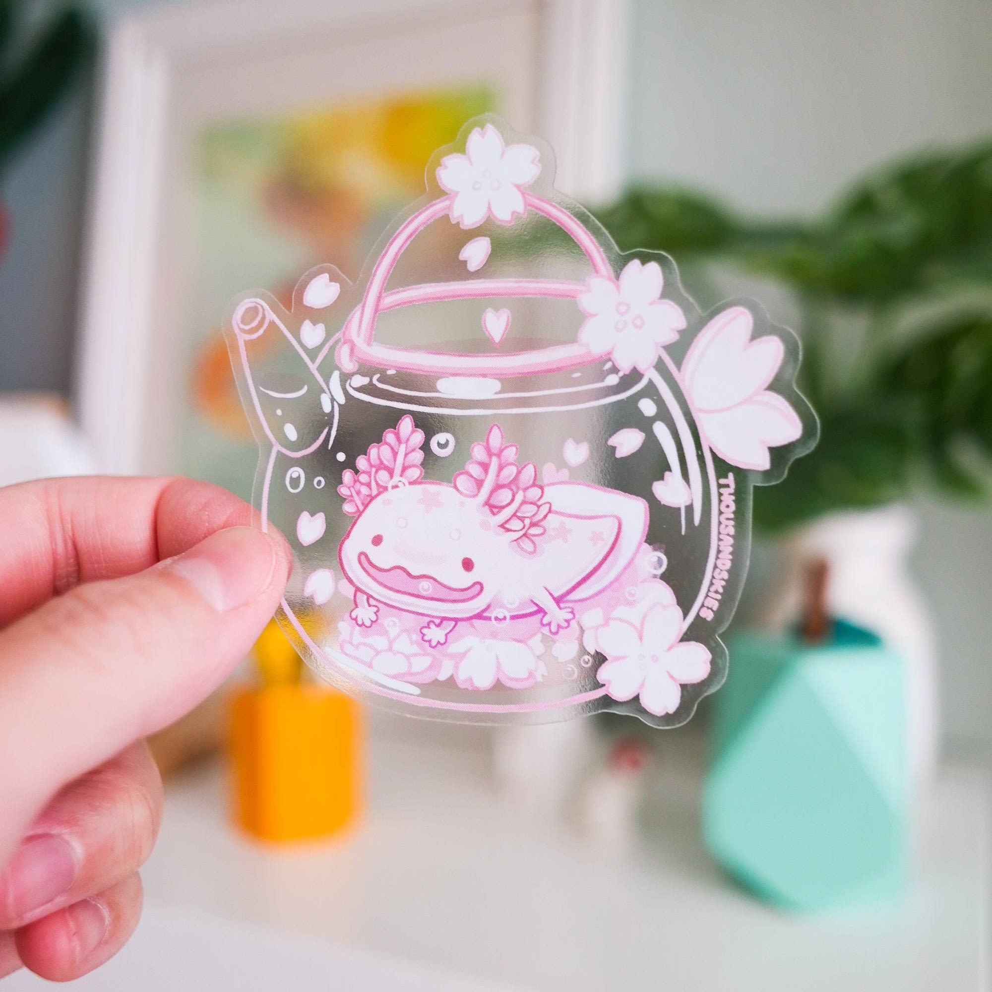 Hand holding a clear acrylic sticker with a pink axolotl design on a blurred indoor background