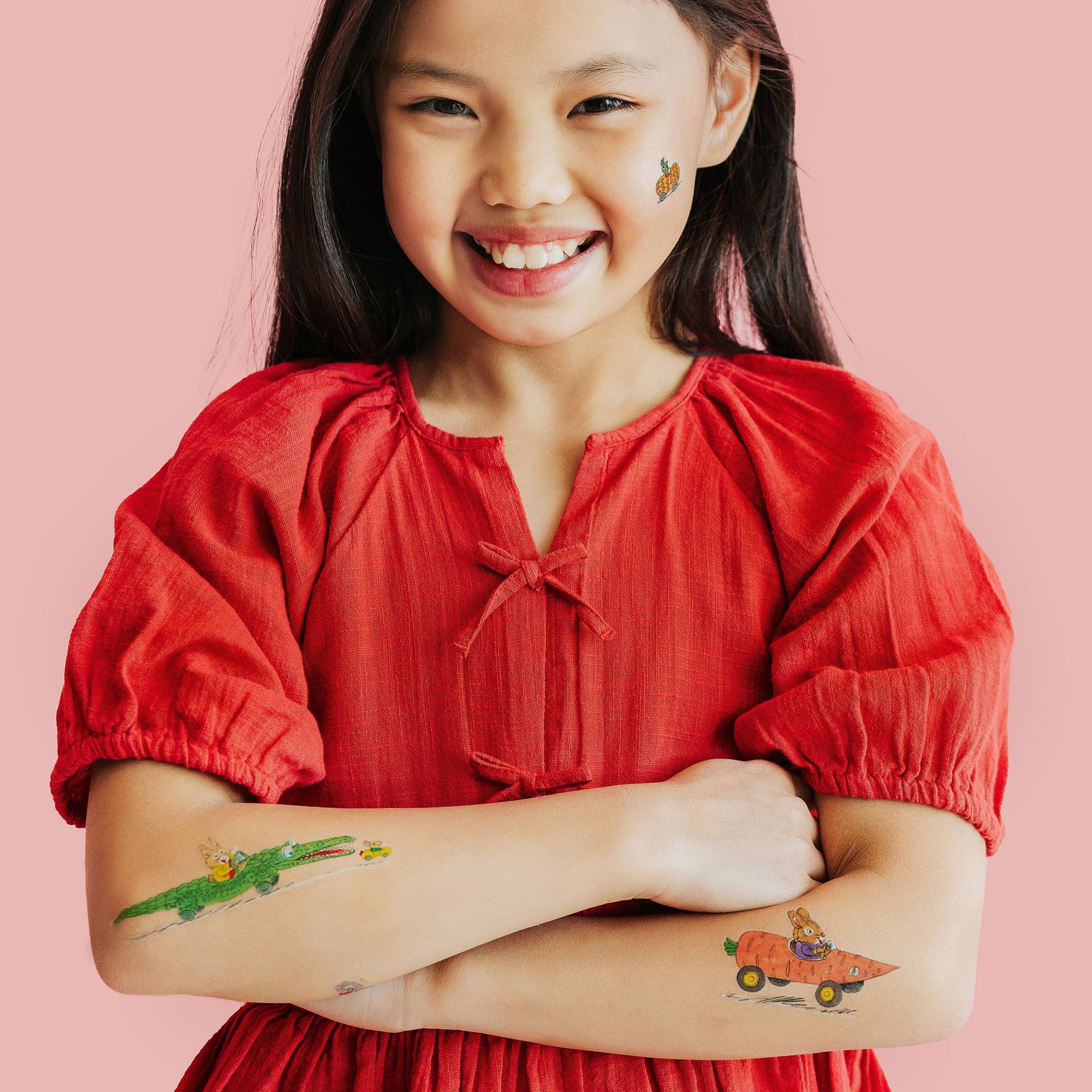Young girl wearing a red blouse with tattoos on her arms against a pink background