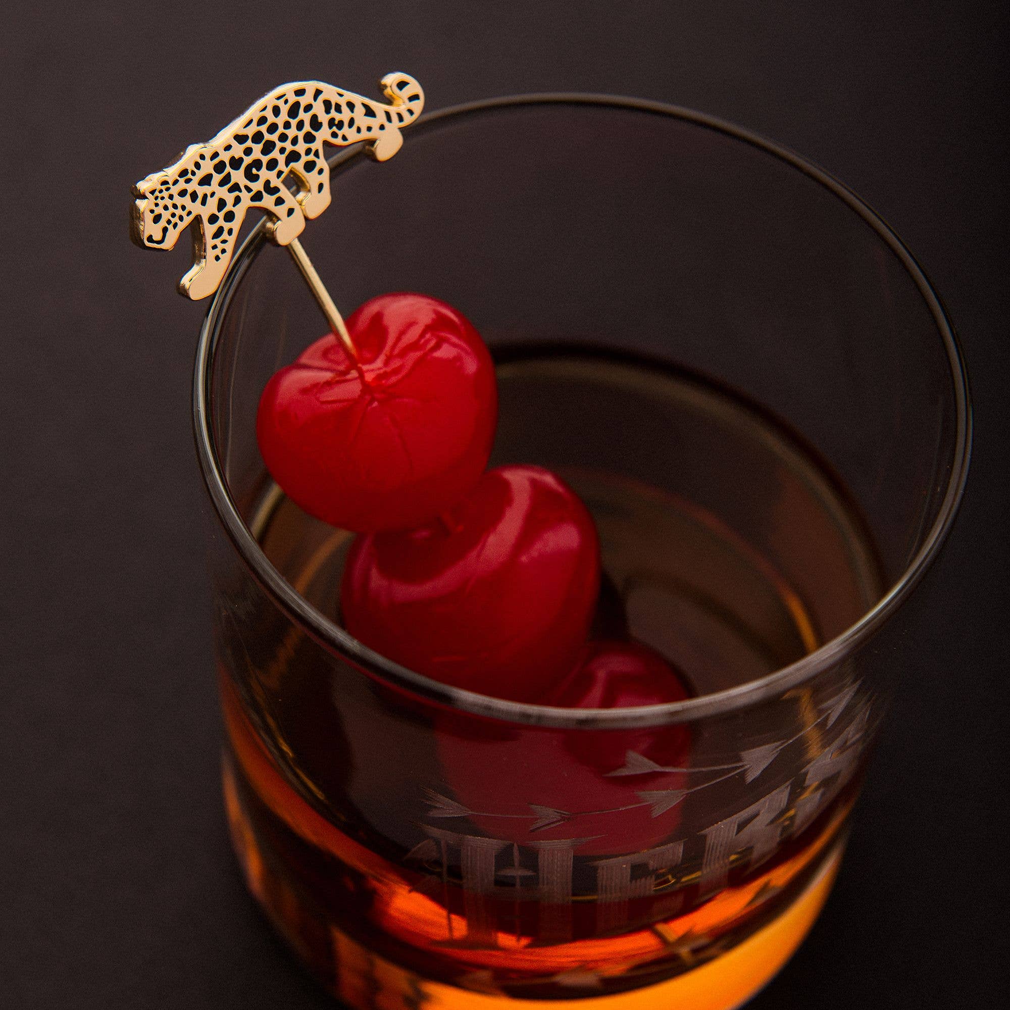 Glass of whiskey with cherries and a gold cocktail pick on a dark background