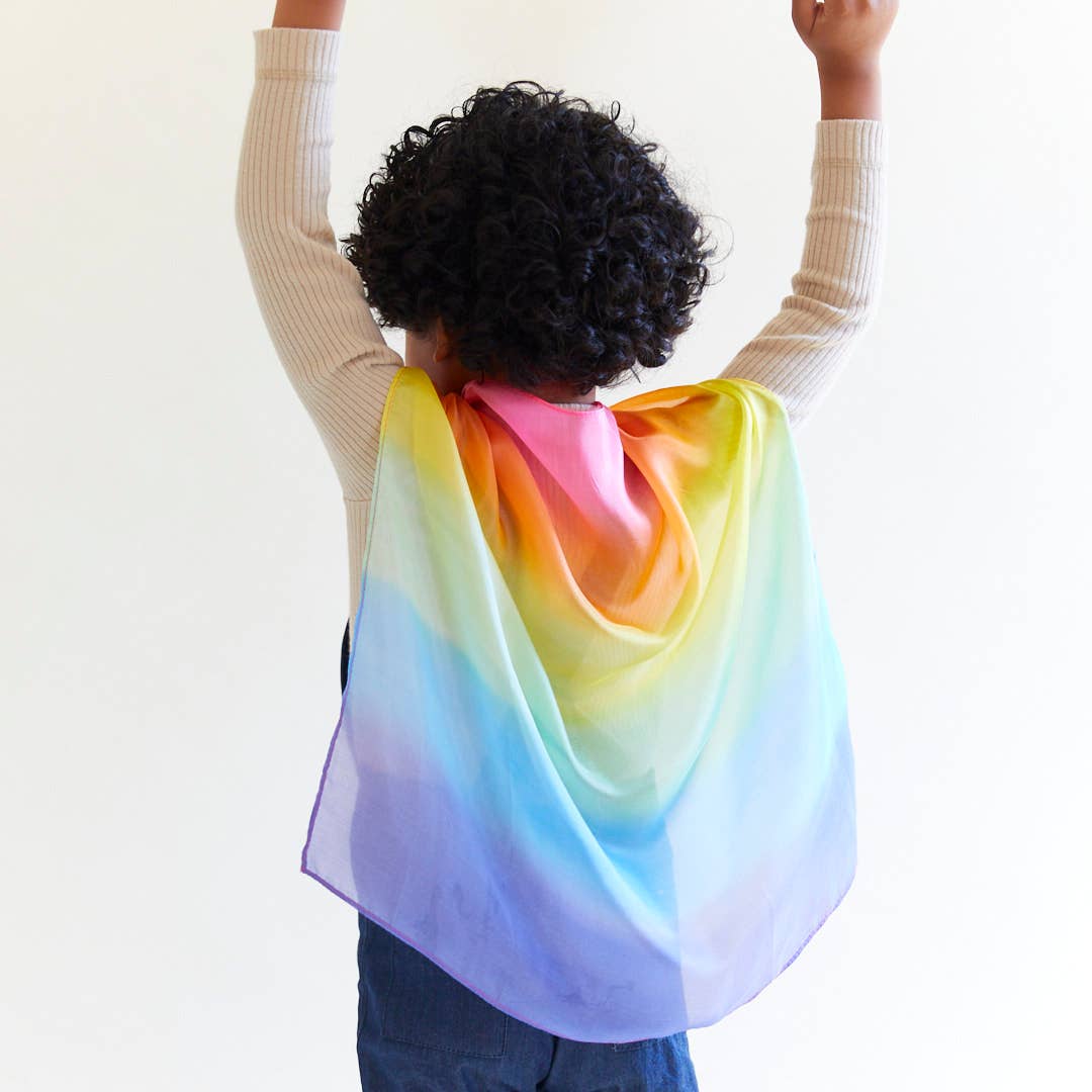 Child wearing a colorful rainbow cape with arms raised against a white background