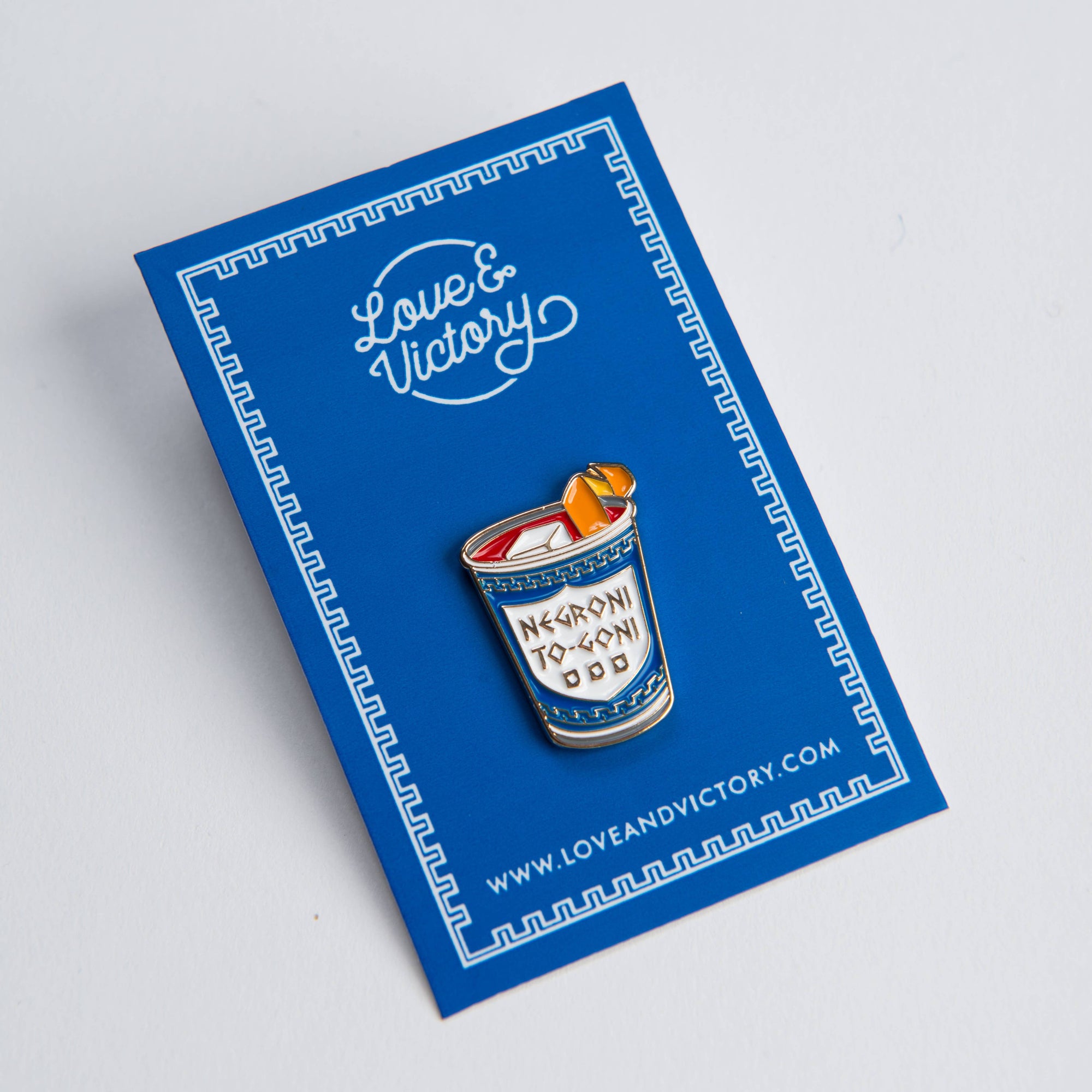 Enamel pin of a cocktail on a blue card with 'Love & Victory' branding.