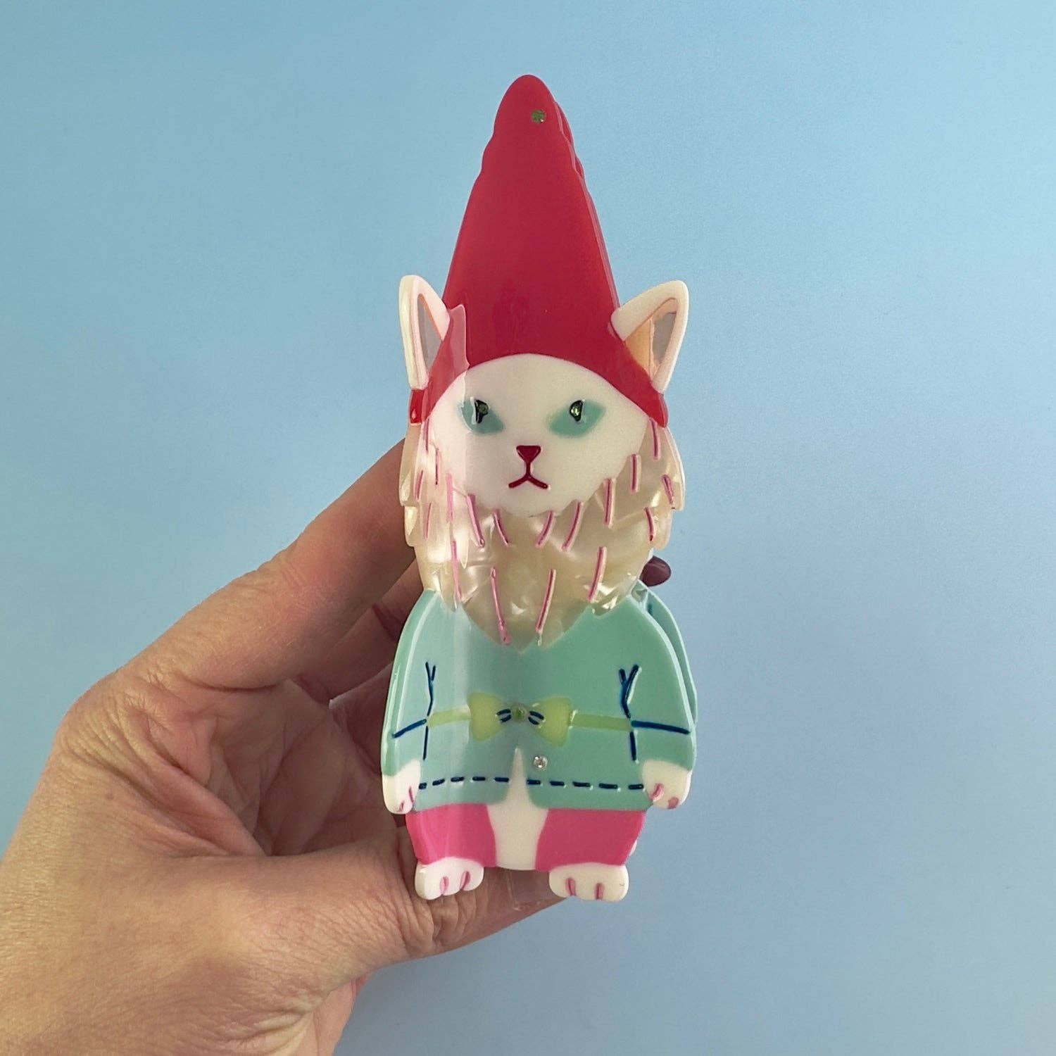 Hand holding a cat-shaped gnome figurine with a red hat against a light blue background