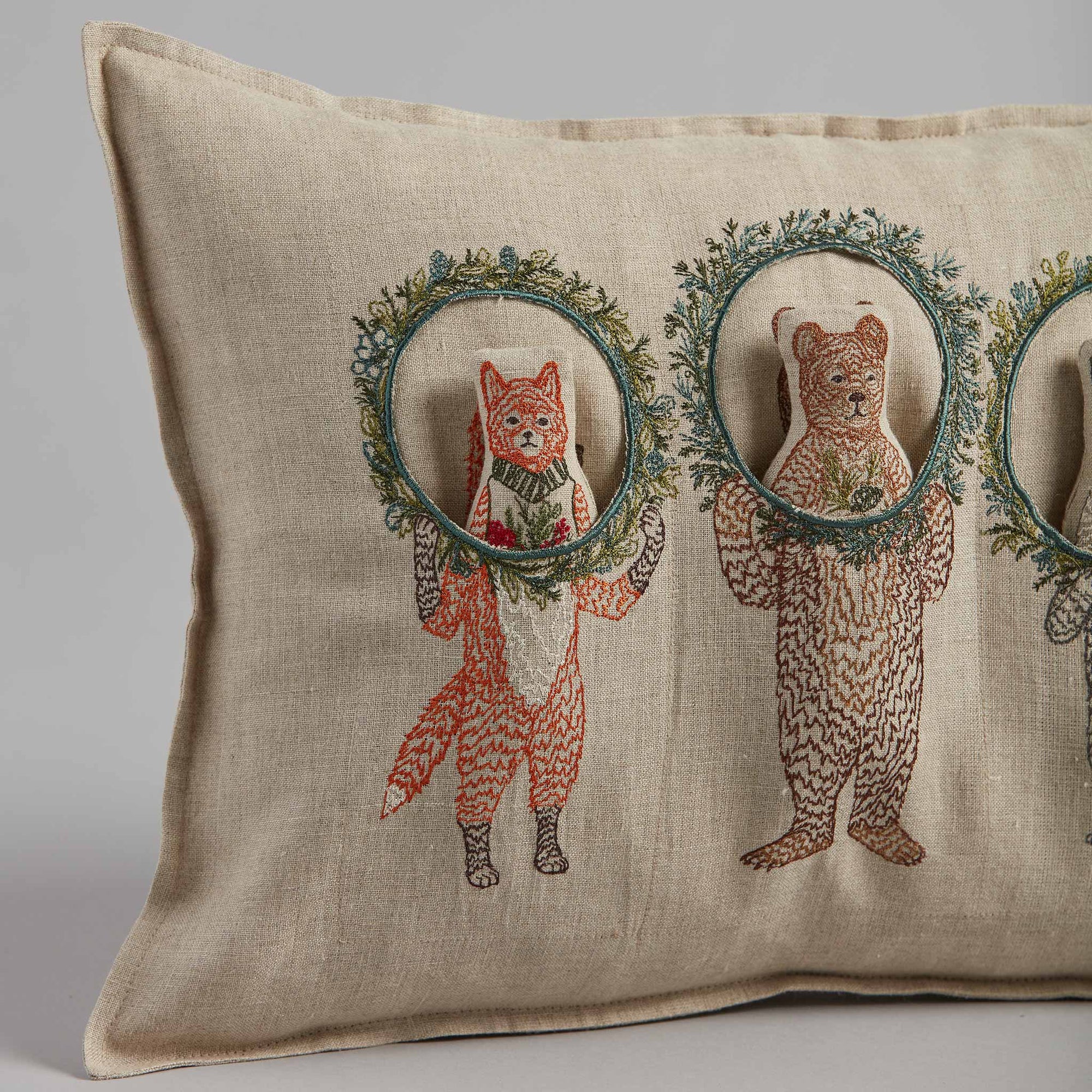 Decorative pillow with embroidered fox and bear holding wreaths on a beige background