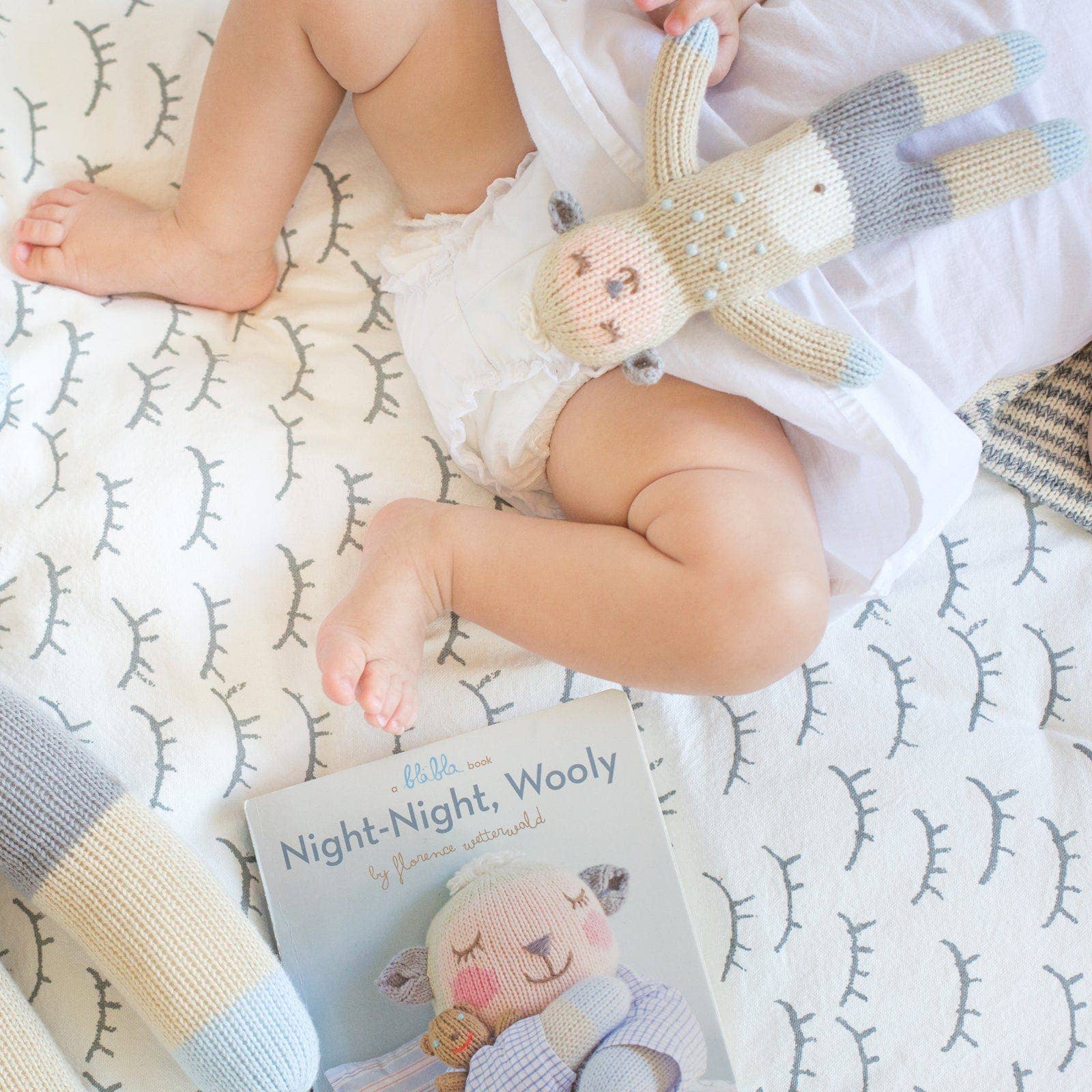 Baby lying on a patterned blanket with a knitted toy and a book titled 'Night-Night, Wooly'.
