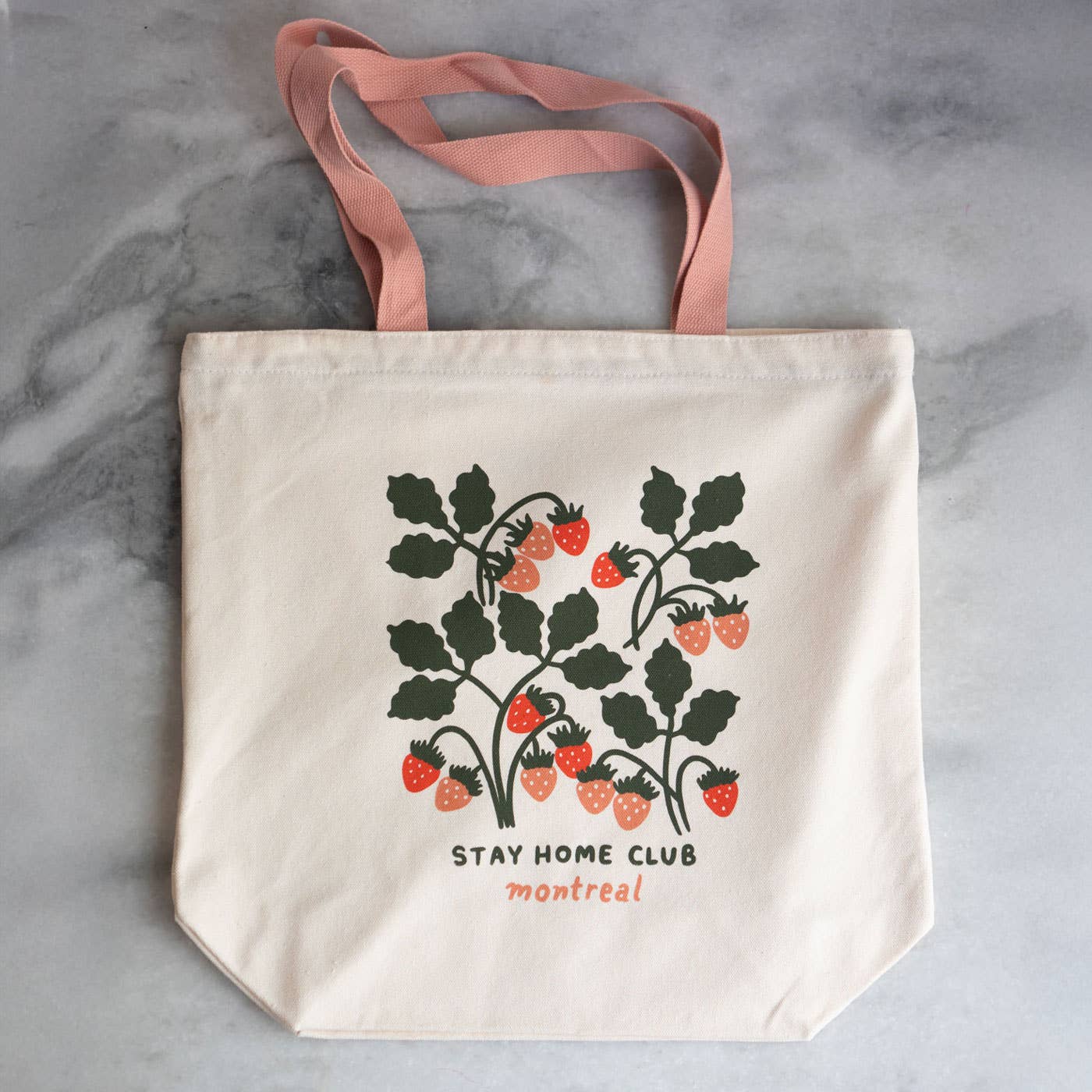 A beige cotton canvas tote bag with pink straps, featuring a vintage-inspired strawberry design and the text 'STAY HOME CLUB montreal'.