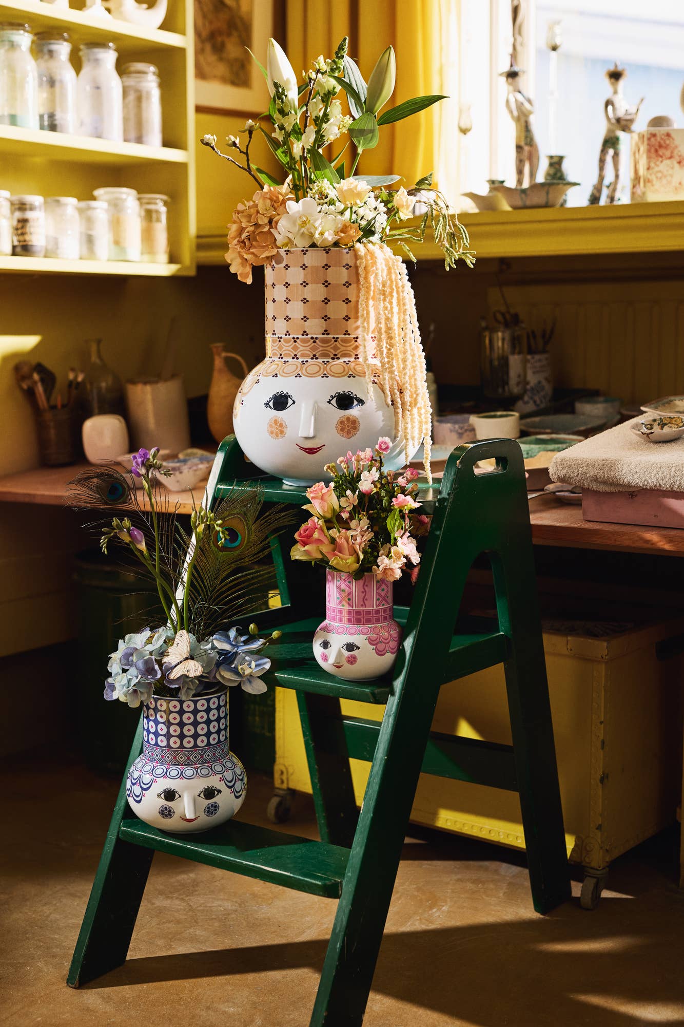 Decorative scene with a small ladder, doll head, and floral arrangements in a room.
