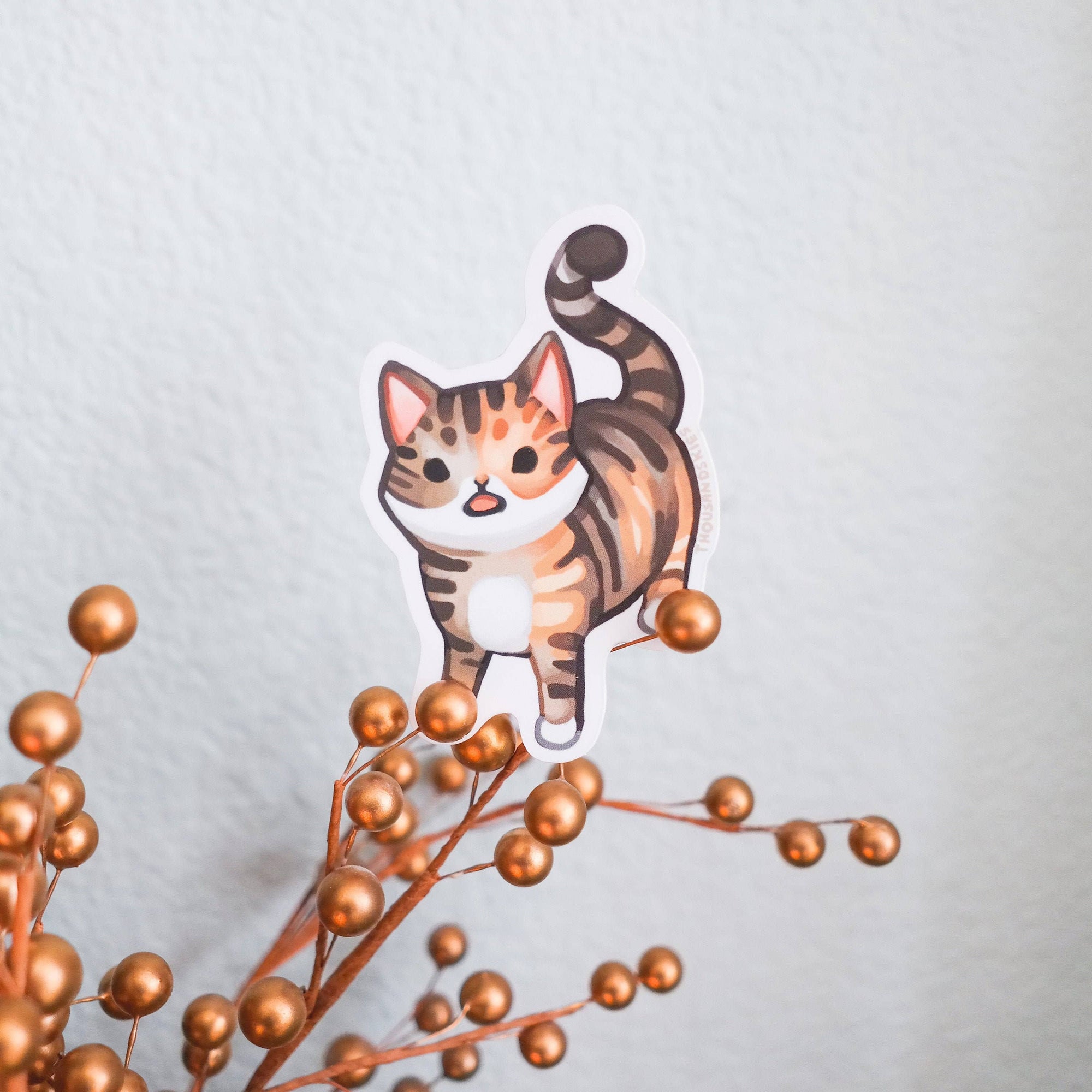 Sticker of a tabby cat on a branch with small gold balls against a light gray background