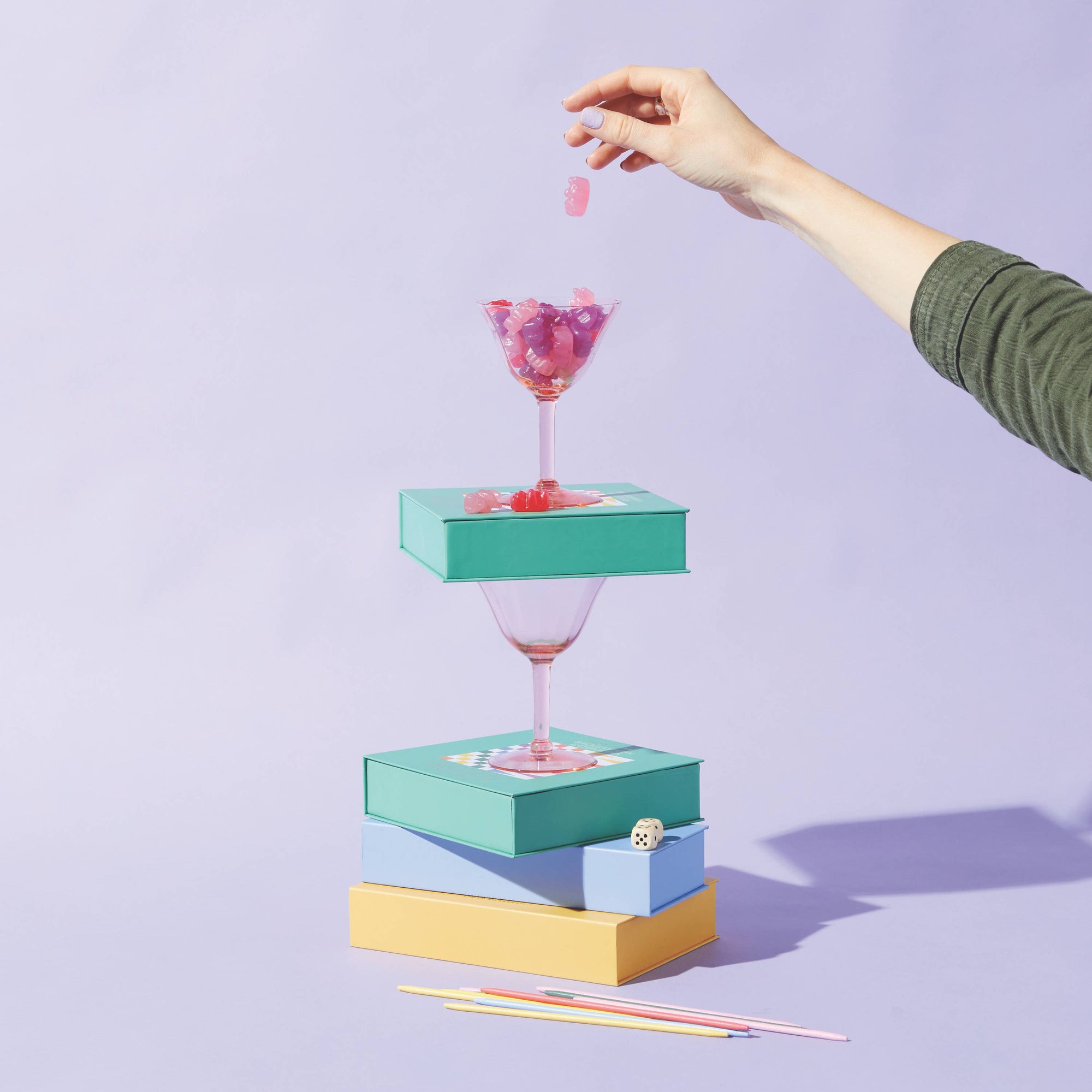 Hand holding a colorful cocktail above stacked gift boxes on a light purple background