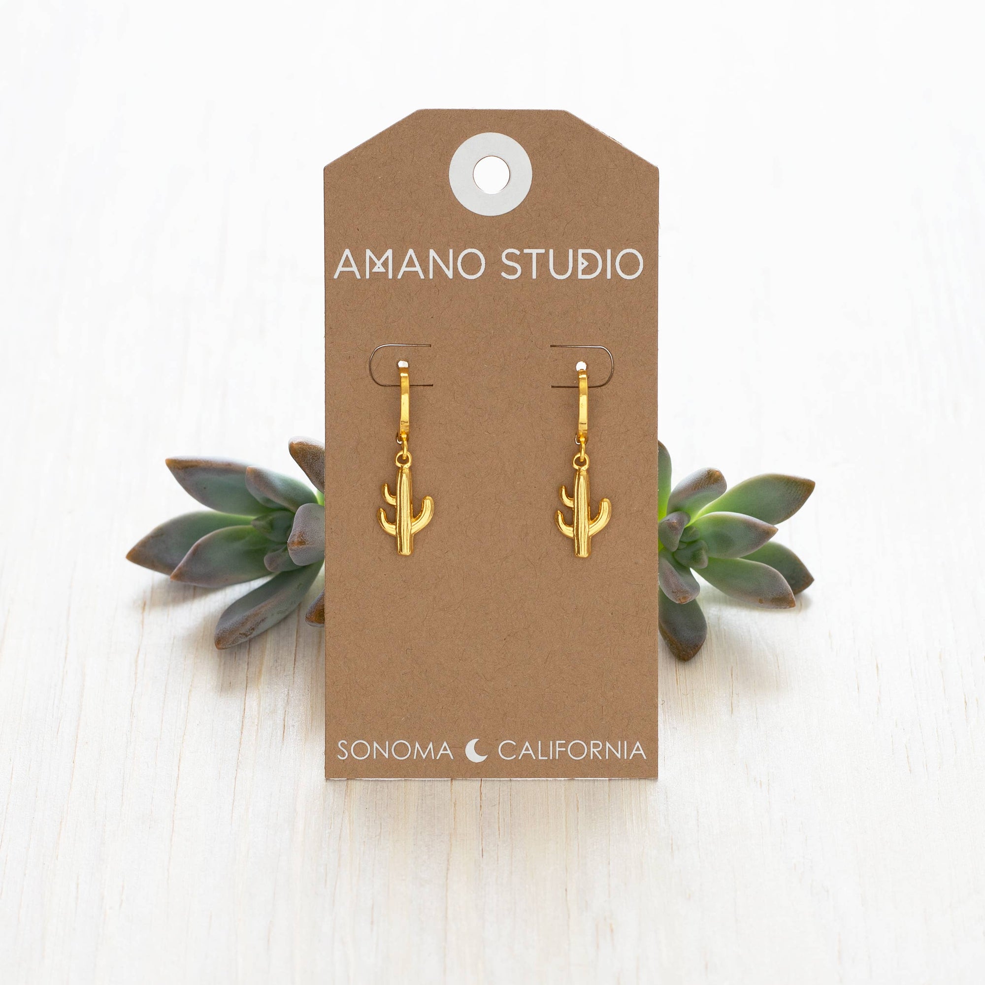 Gold cactus-shaped earrings on a 'Amano Studio' branded card with succulents in the background.