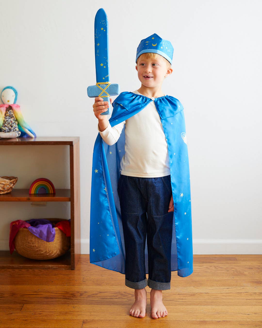 Child in a blue superhero costume holding a toy sword in a room with a shelf in the background.