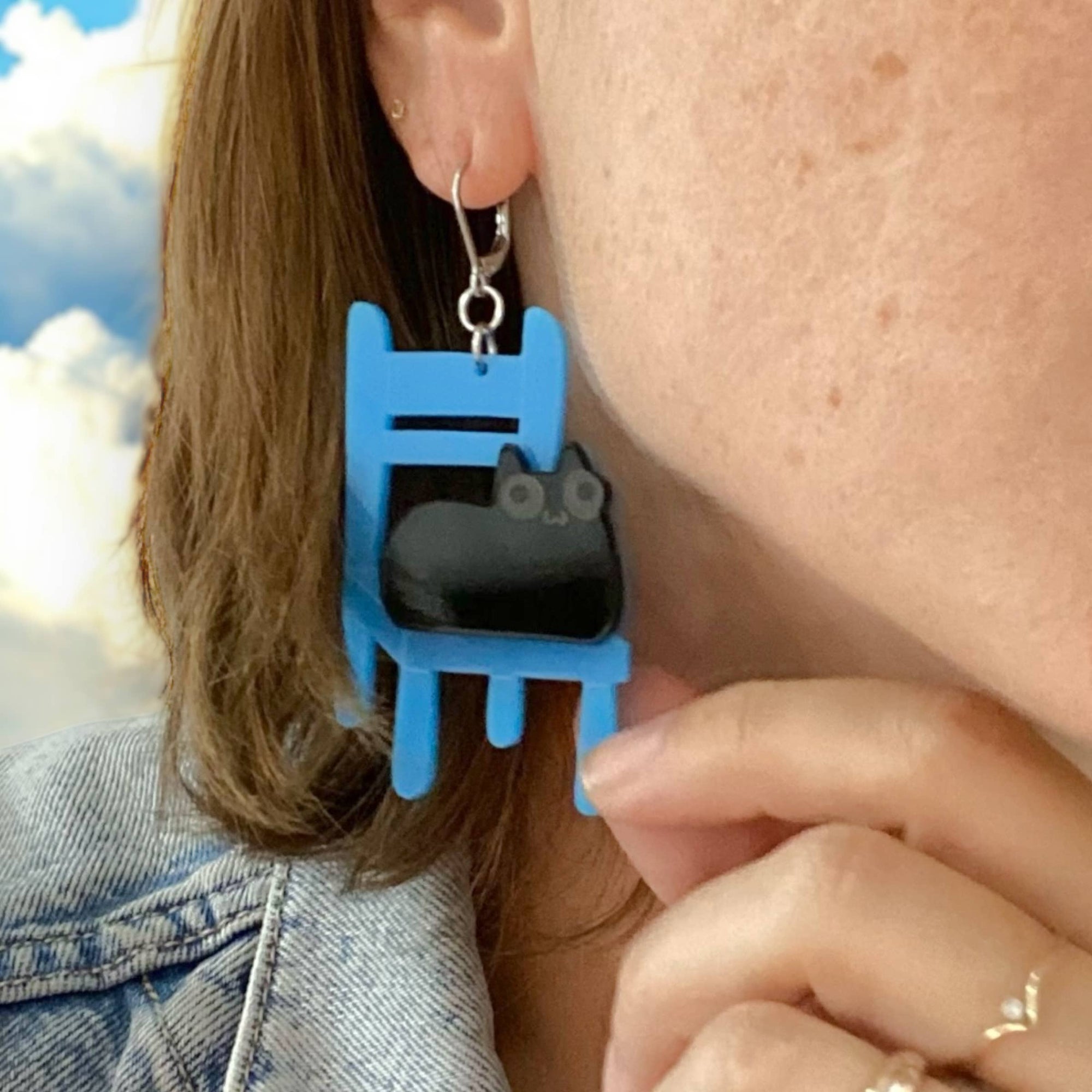 Person wearing blue and black cat-shaped earrings with a blurred outdoor background