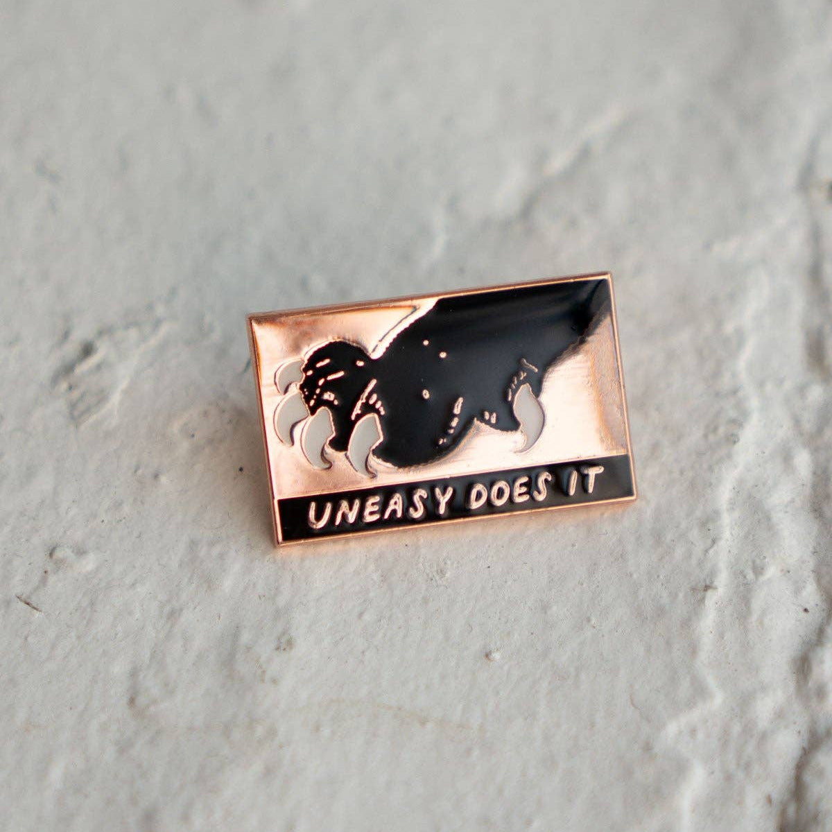 Rectangular pin with a black silhouette and 'Uneasy Does It' text on a gray surface