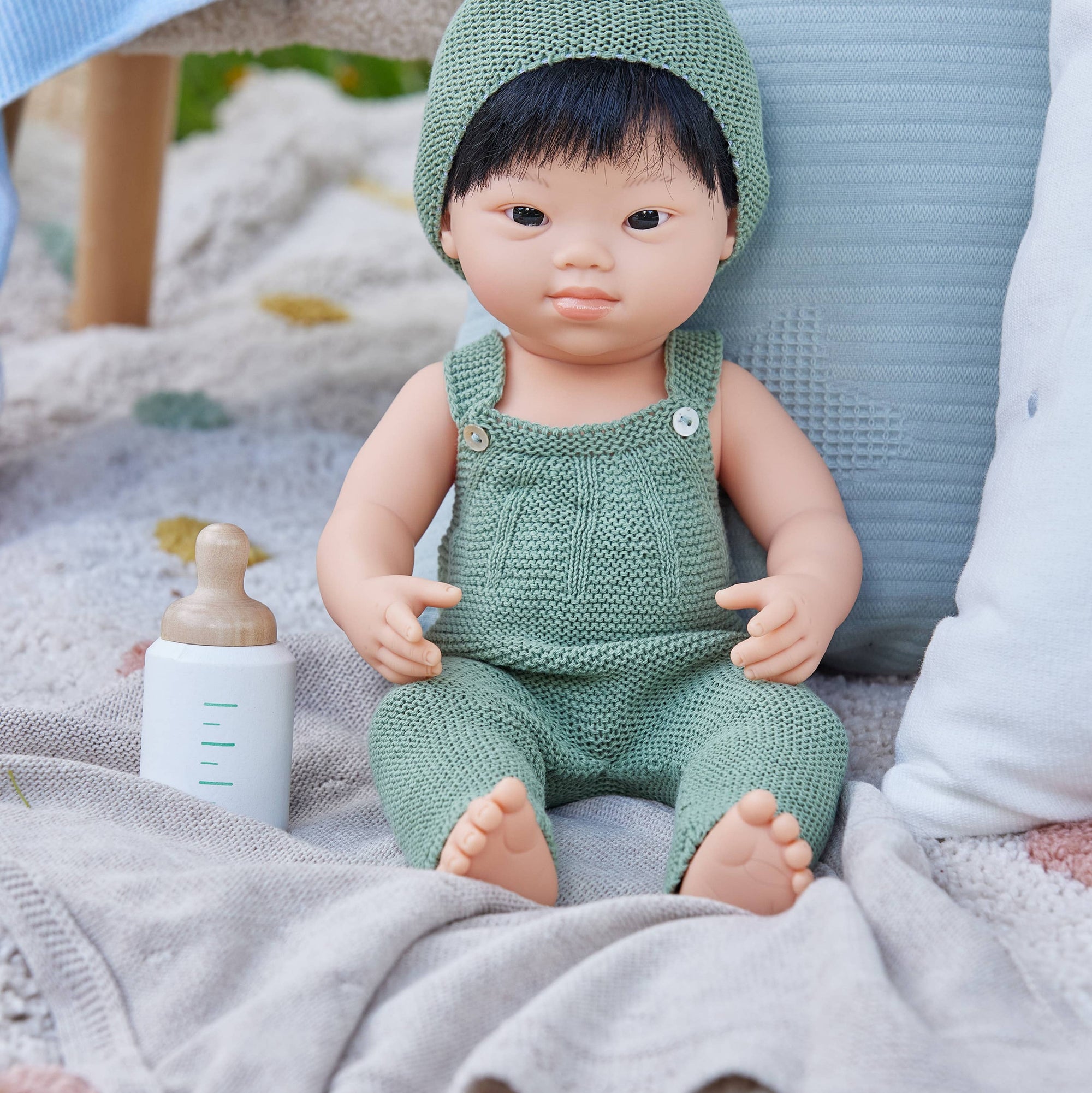 Baby doll in a green outfit sitting on a blanket with a bottle and toy in the background.