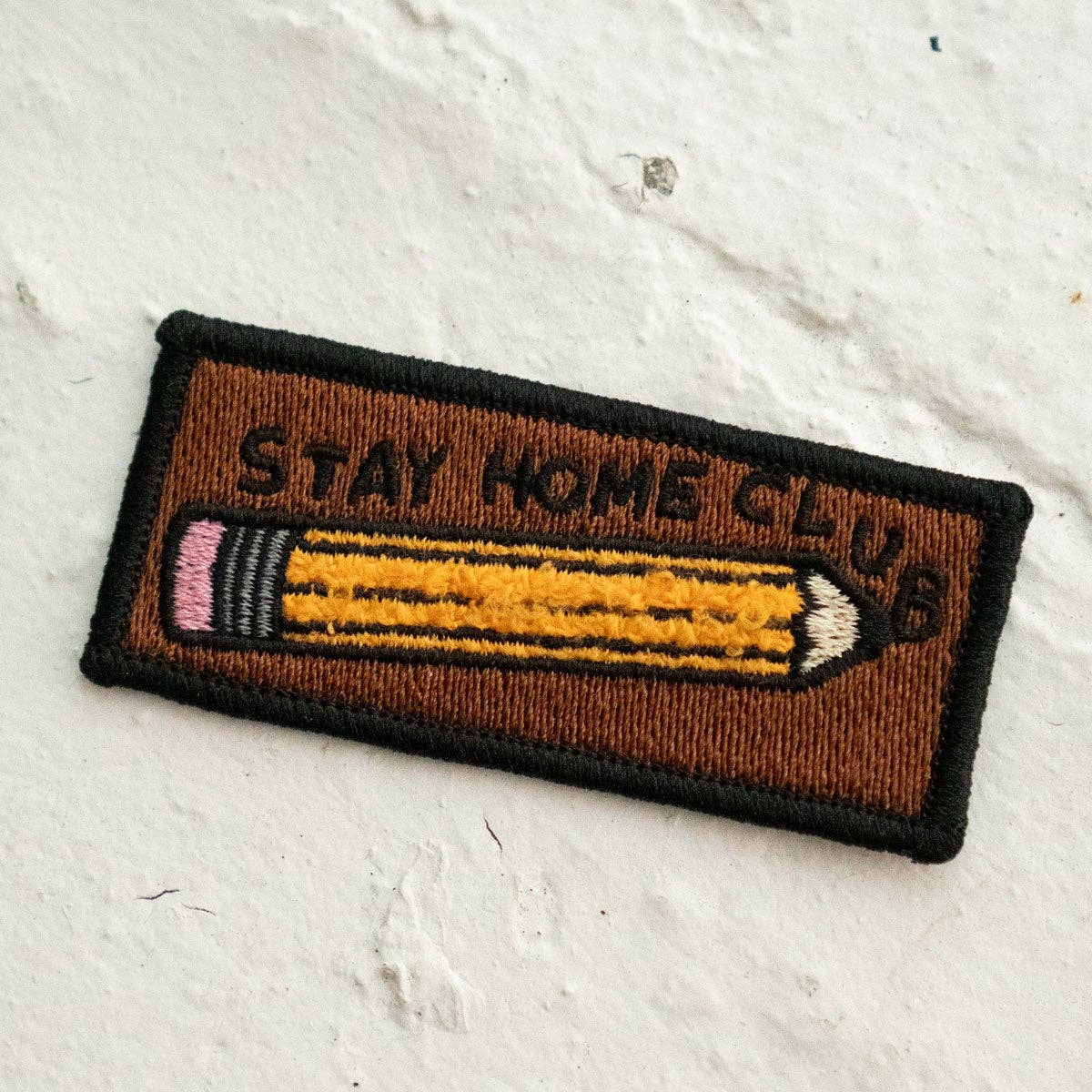 Embroidered patch with a pencil design and 'Stay Home Club' text on a textured white background