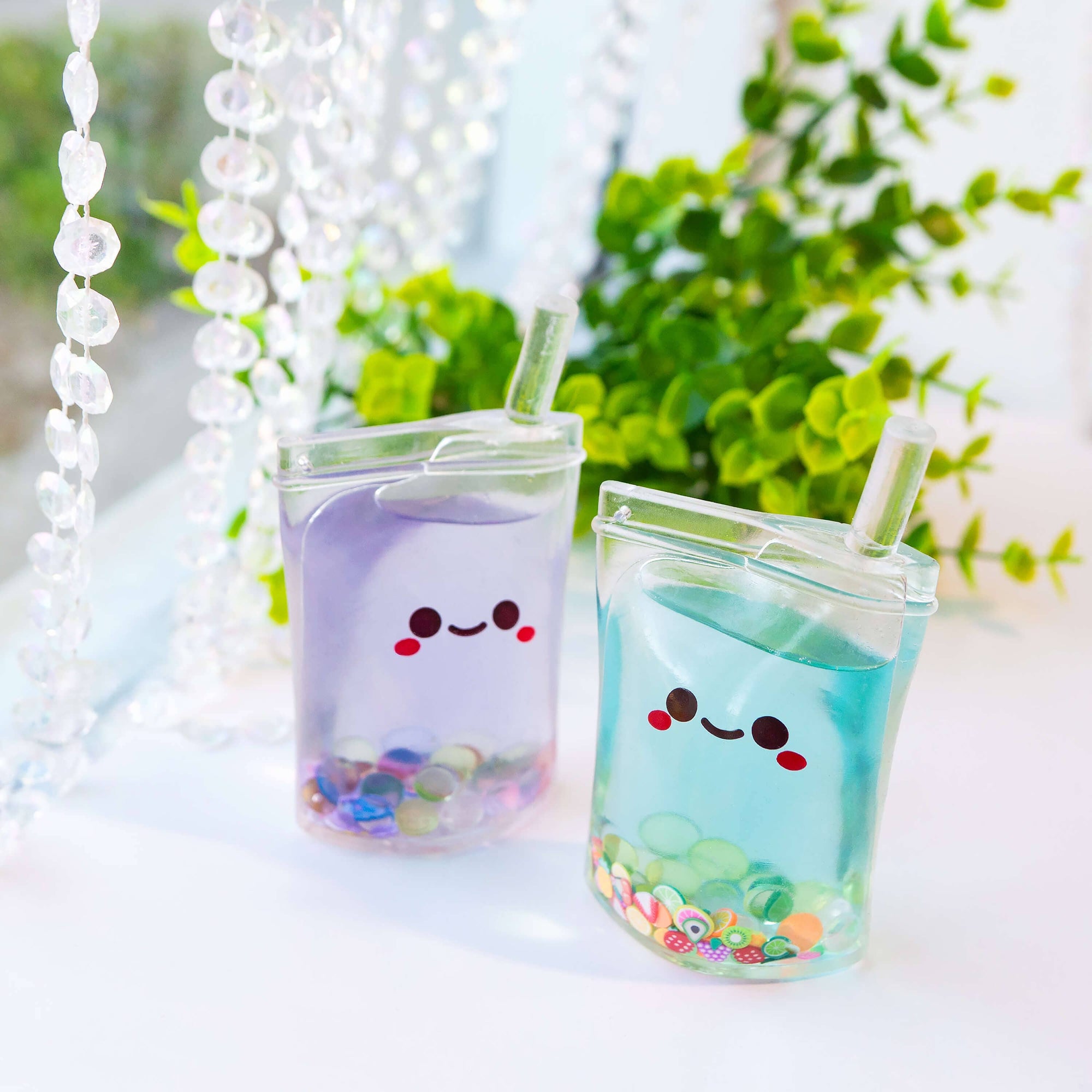Two colorful drink containers with smiling faces on a light surface with greenery in the background.