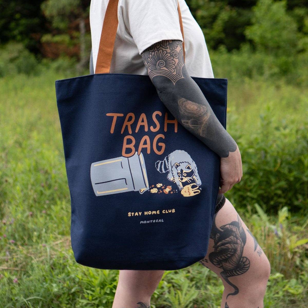 Person holding a navy tote bag with 'Trash Bag' text and graphic, outdoors.