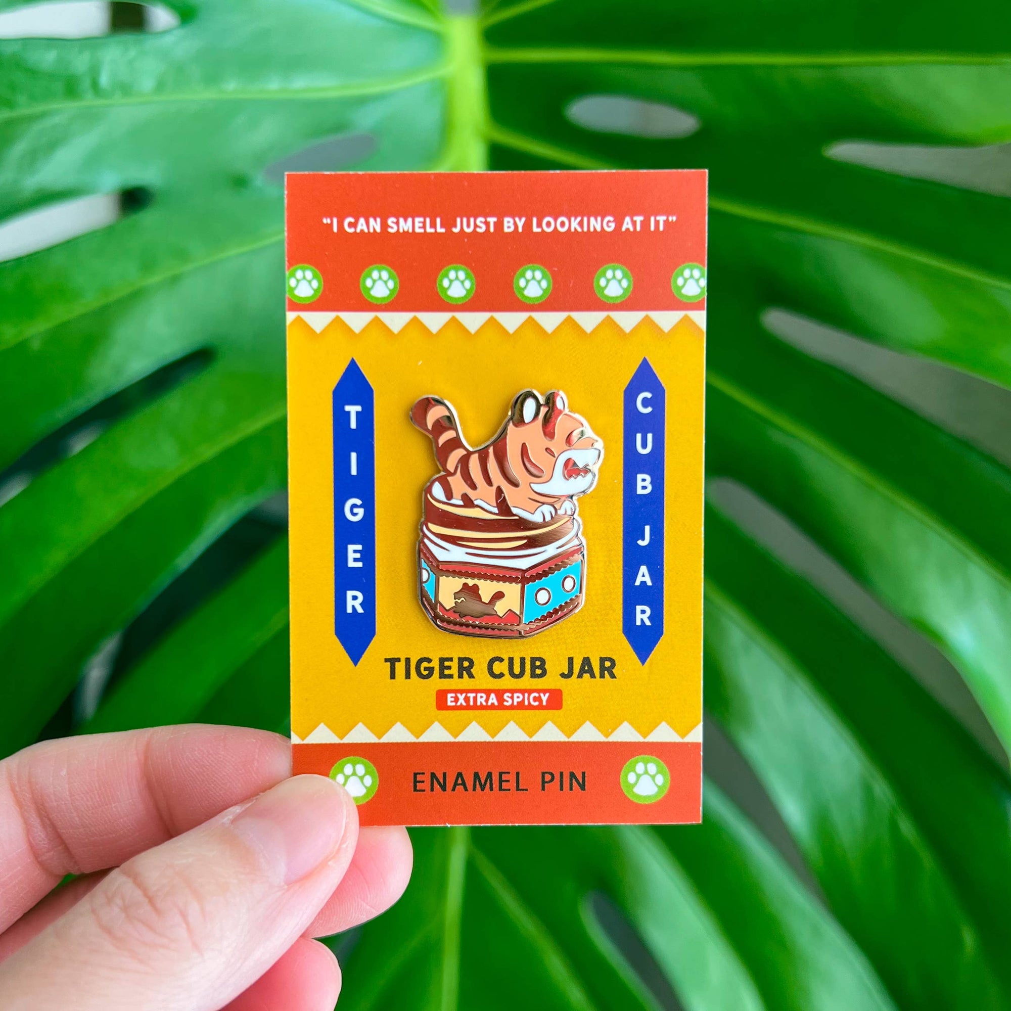 Enamel pin of a tiger cub jar with a colorful label against a green leaf background