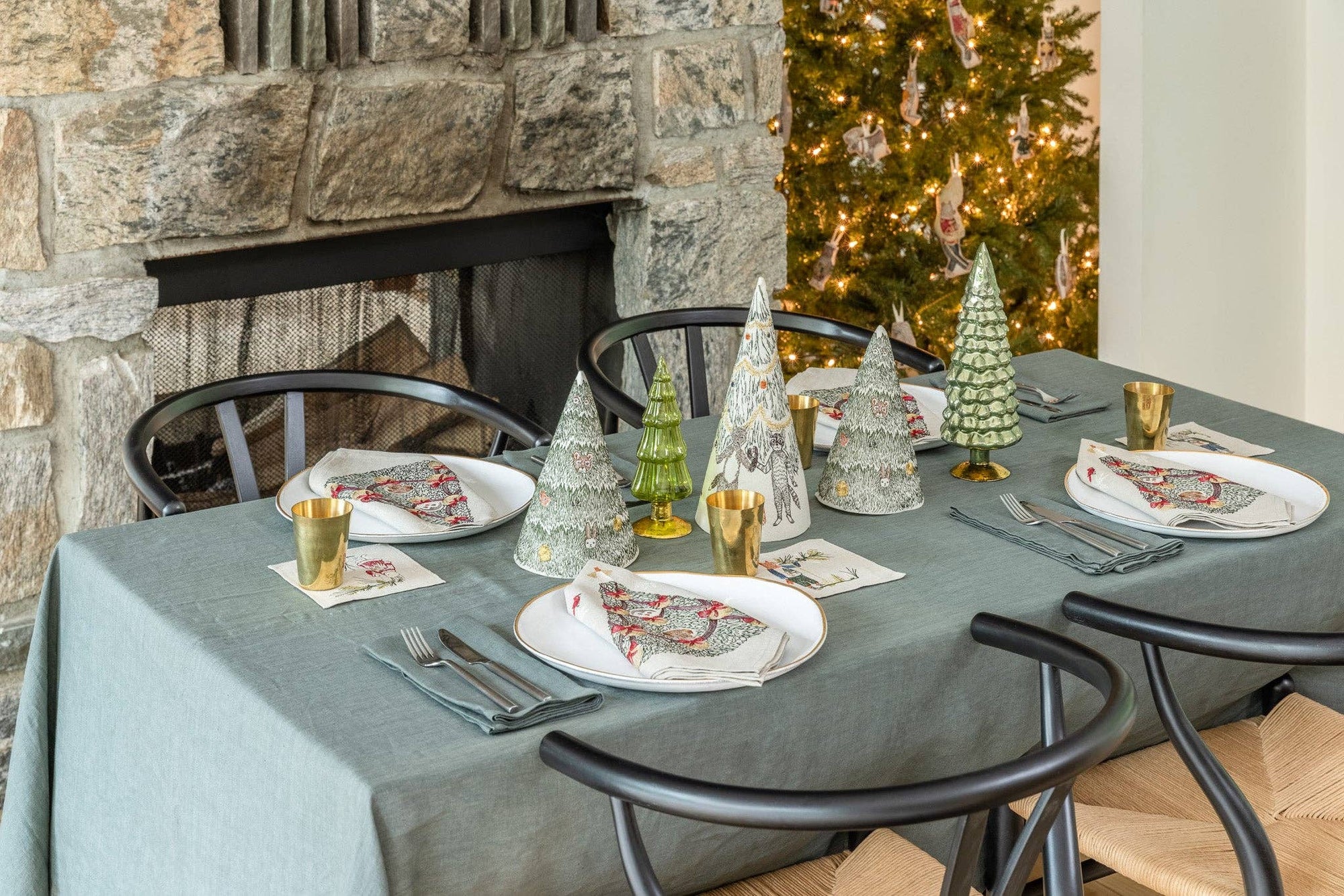 Dining table set for Christmas with decorative trees and place settings in front of a stone fireplace.