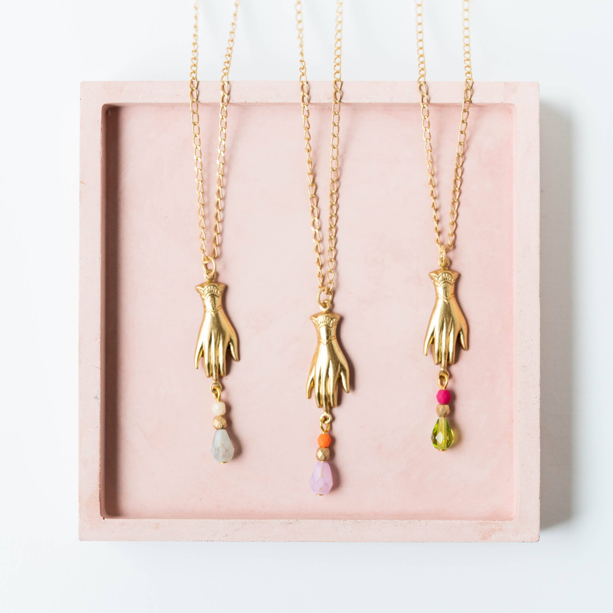 Three gold hand-shaped pendants on chains with colorful beads in a pink display box.