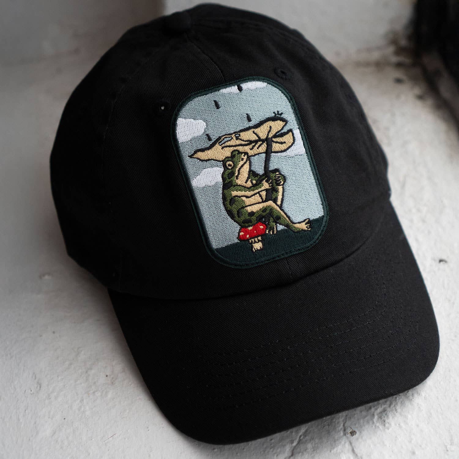 Black cap with a embroidered design of a bear holding an umbrella on a white surface