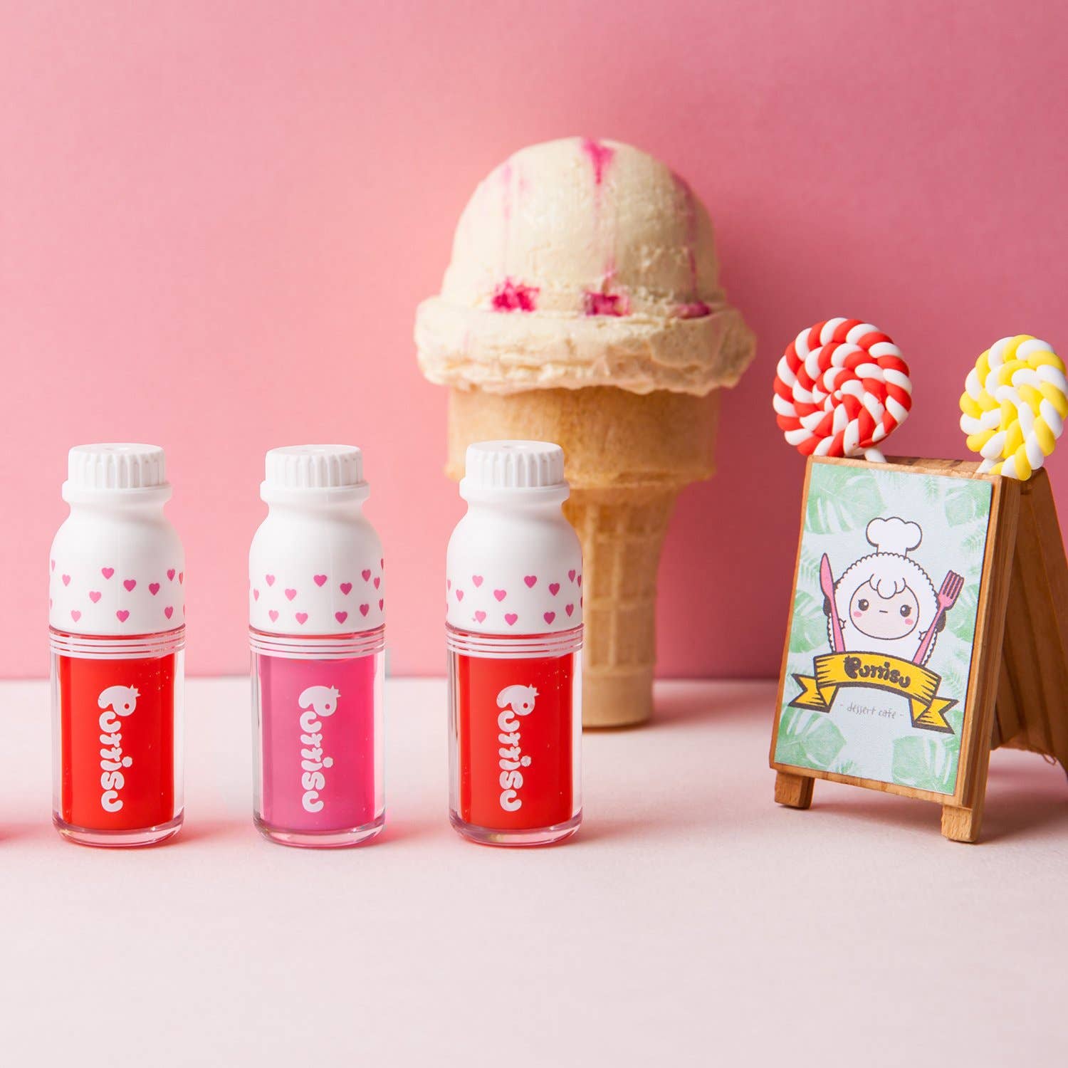 Three small bottles of lip gloss with red and pink designs next to an ice cream cone on a pink background