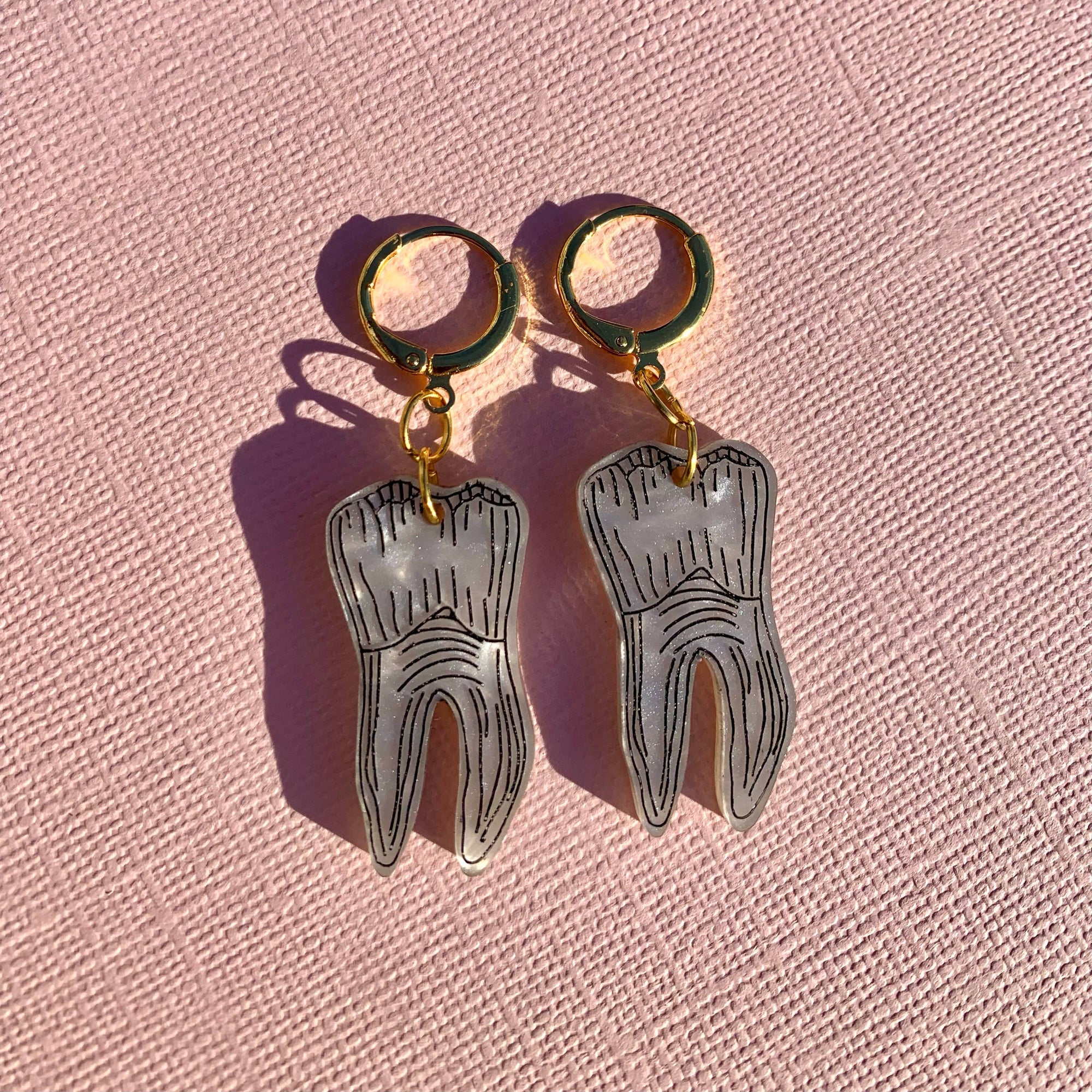 Silver tooth-shaped earrings on gold hooks against a pink textured background