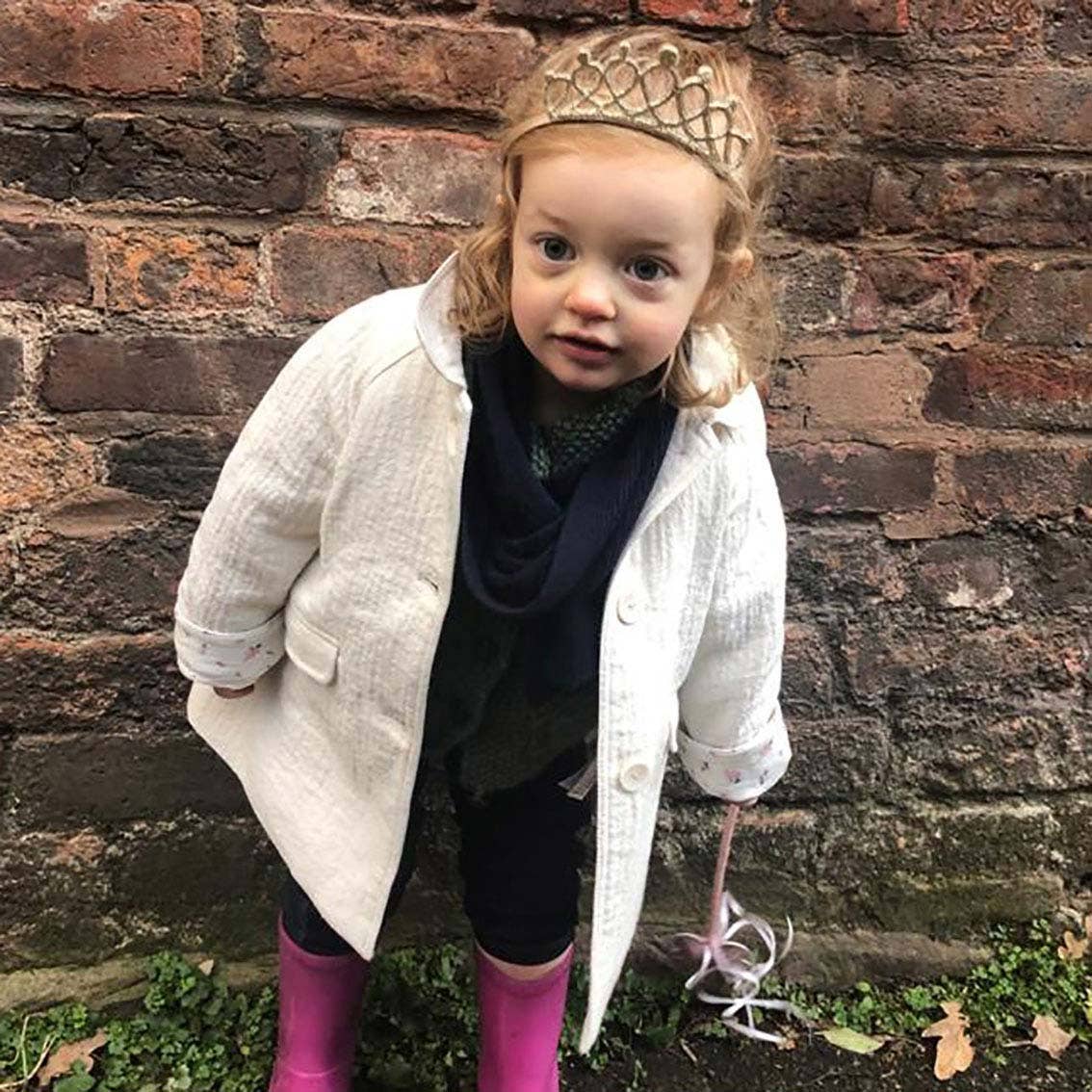 Child wearing a white coat, black dress, pink boots, and a gold tiara against a brick wall.