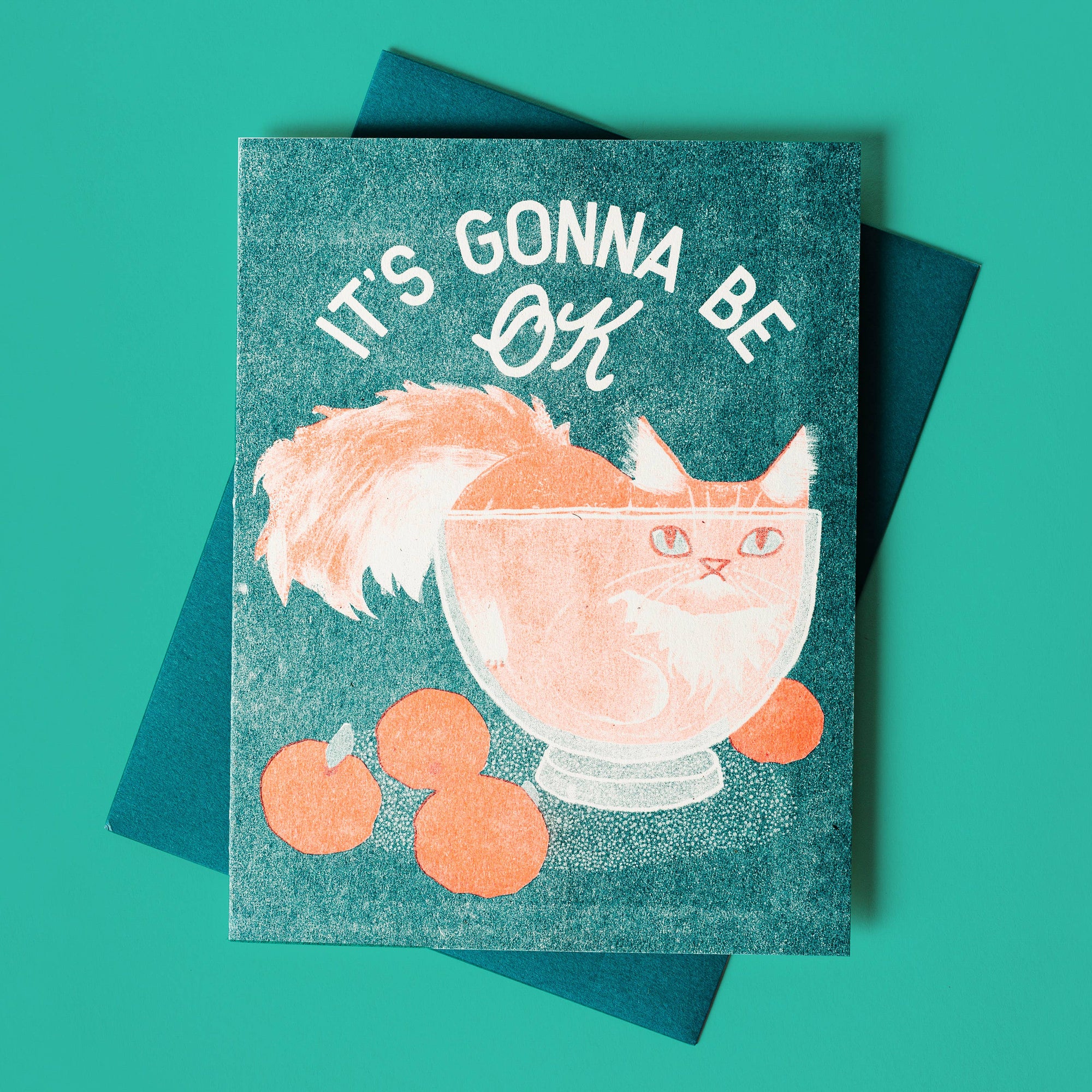 A Greeting card with an illustration of a cat inside a bowl, surrounded by oranges, with the text 'IT'S GONNA BE OK' above it. The card is placed on a teal background.