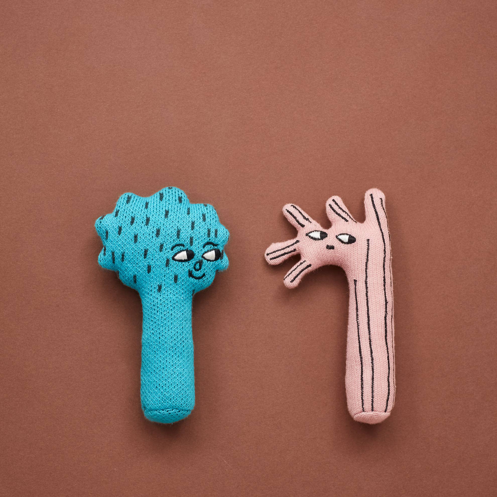 Two knitted baby rattles, one blue with a happy face and raindrop pattern, and one pink with a stick figure design, set against a brown background.