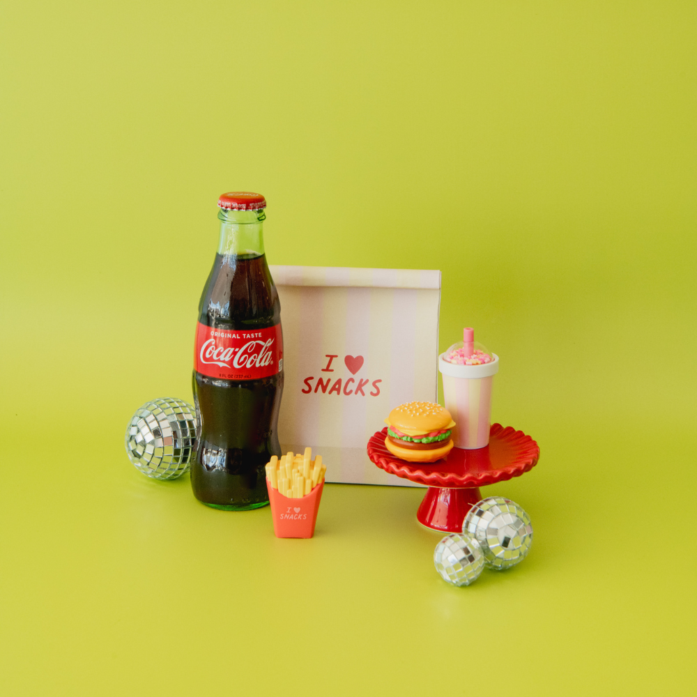 Coca-Cola bottle, snack box with 'I love snacks' text, burger, fries, and pink drink on a green background