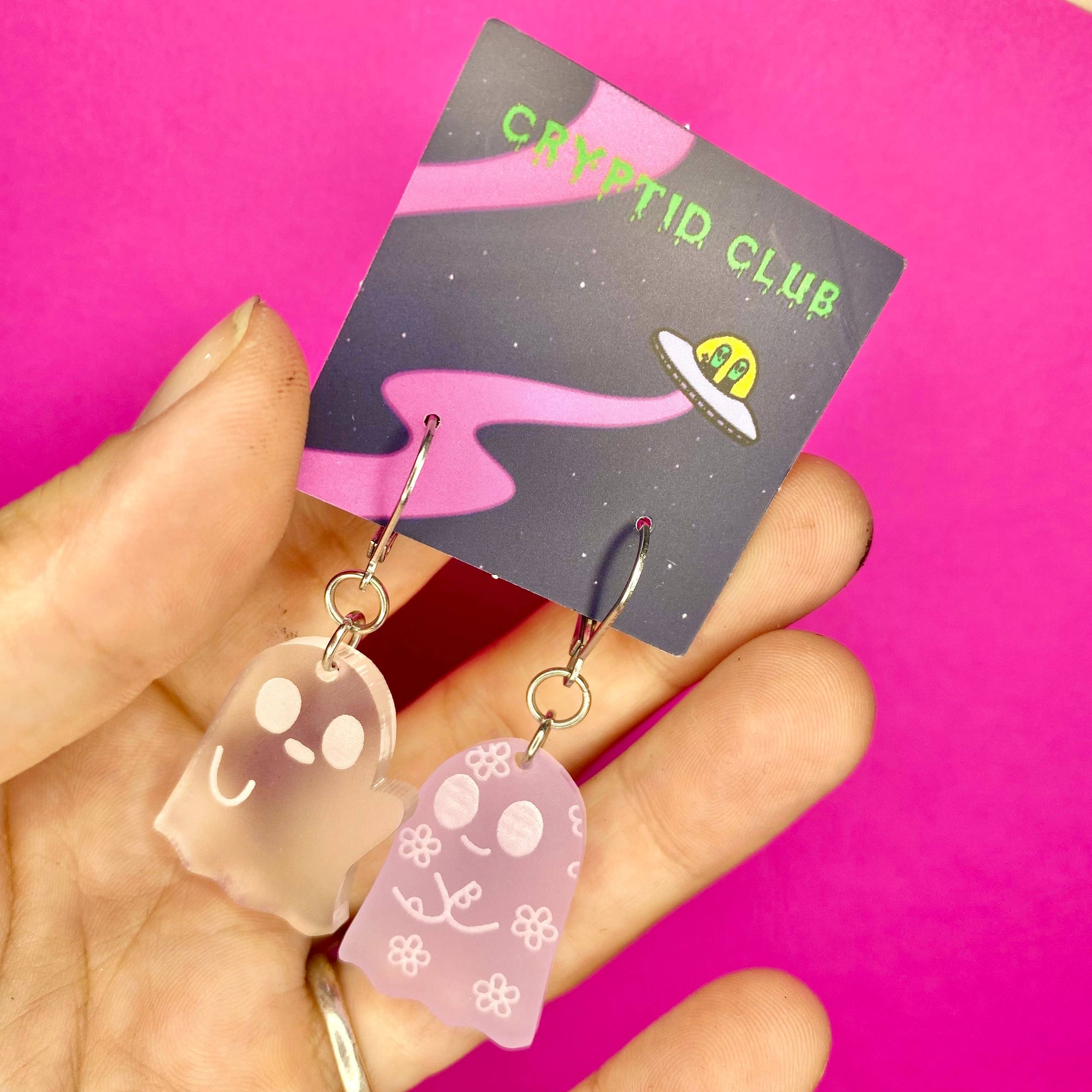 Transparent ghost-shaped earrings held in front of a 'Cryptid Club' card on a pink background