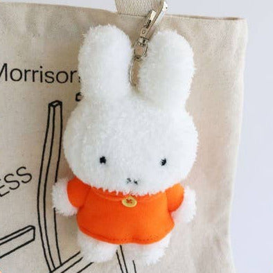 White plush bunny keychain with an orange sweater on a beige bag with text.