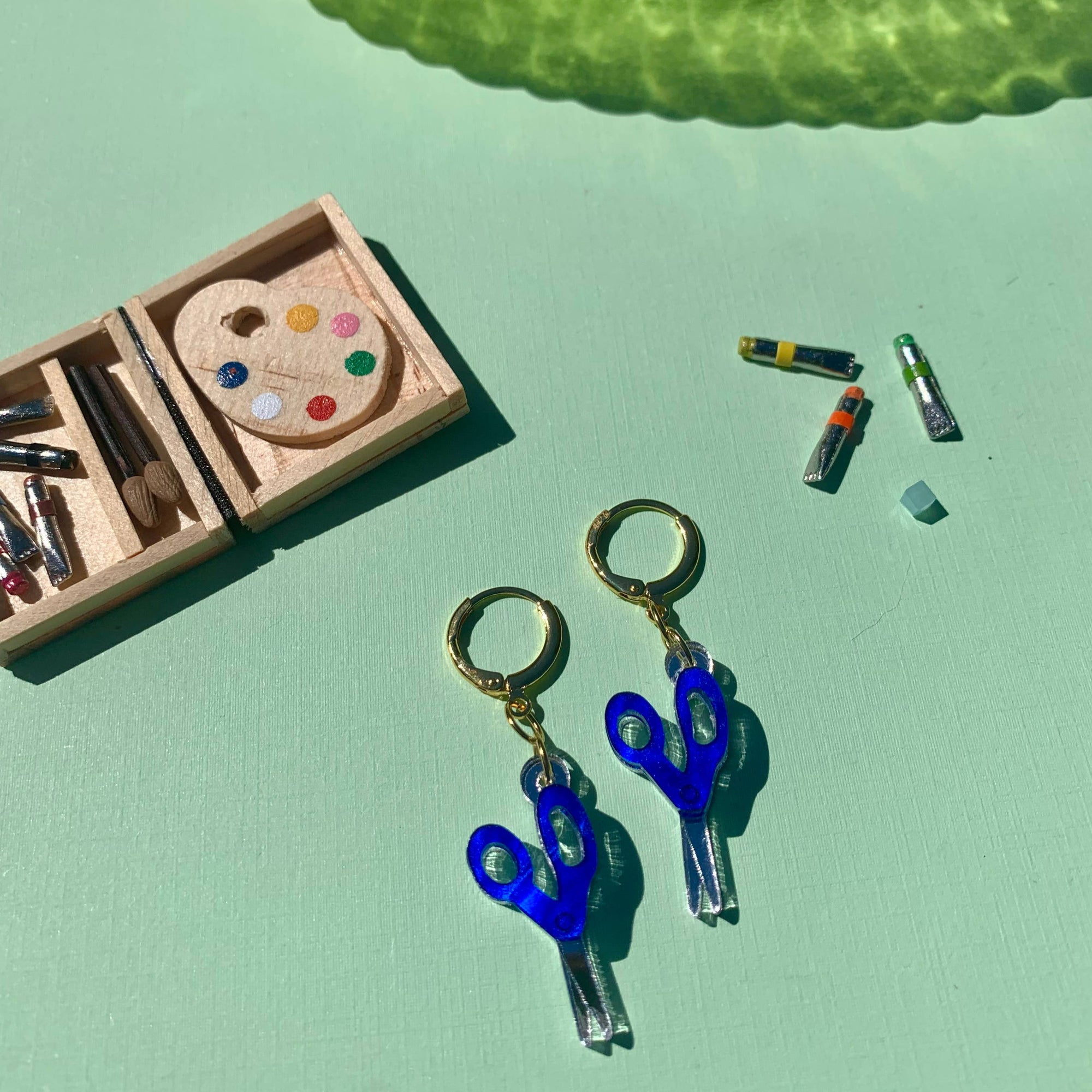 Blue scissors earrings on a light green background with a small wooden palette and markers.