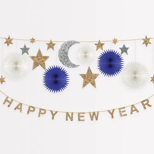 A New Year's garland decoration with a space theme, featuring navy and white honeycomb stars, gold glitter fabric stars and letters, and a silver glitter fabric moon, all strung together with gold metallic cord.