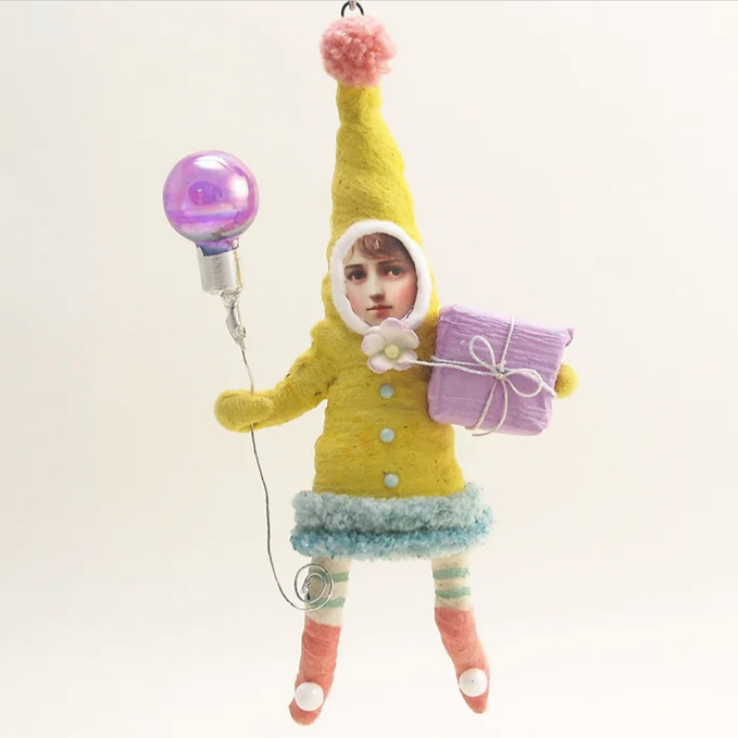 Handmade spun cotton ornament depicting a yellow elf with a striped hatband and blue-green socks, holding a purple balloon and a package, against a white background.