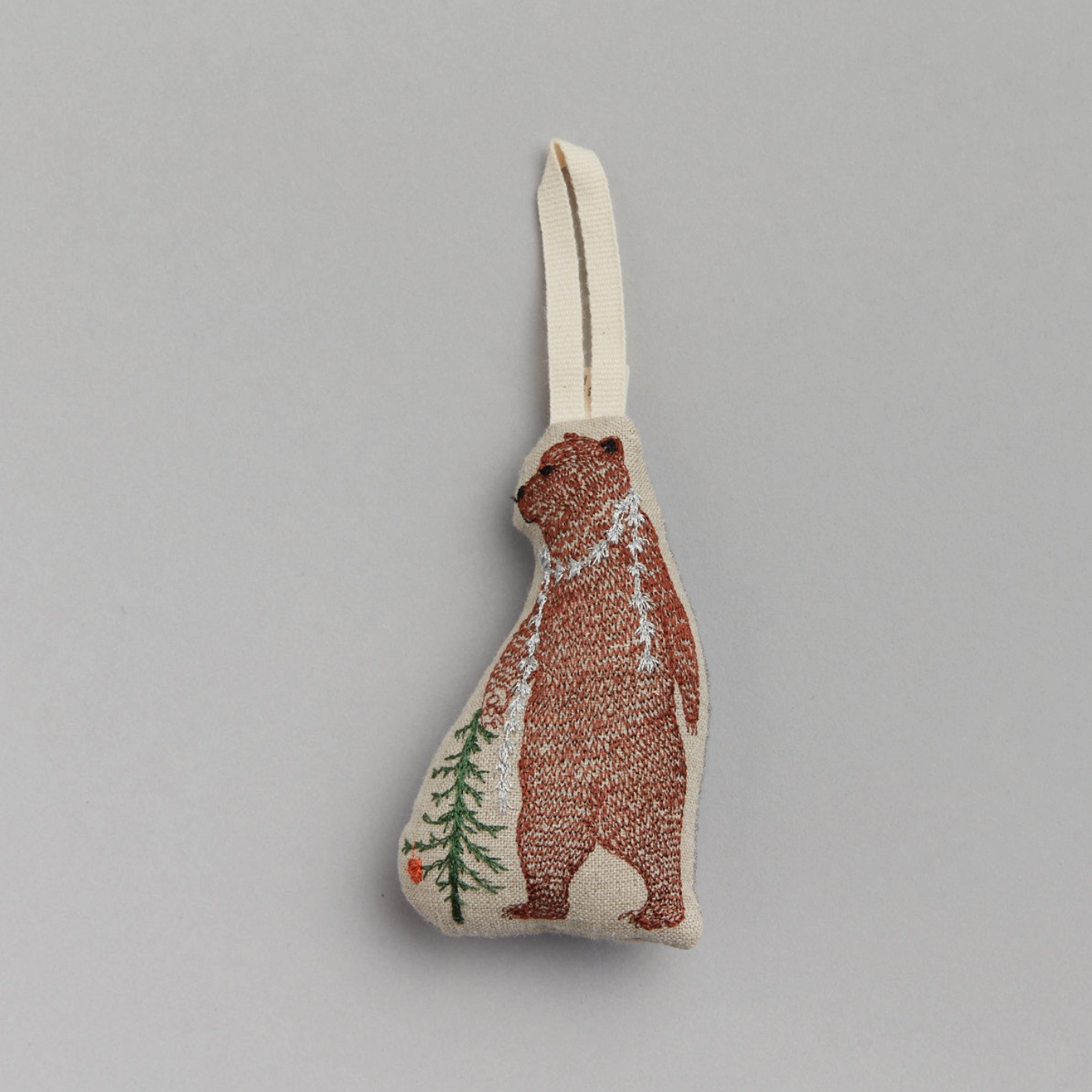 A decorative holiday ornament featuring an embroidered bear with a Christmas tree and tinsel in the background, on a grey linen background.